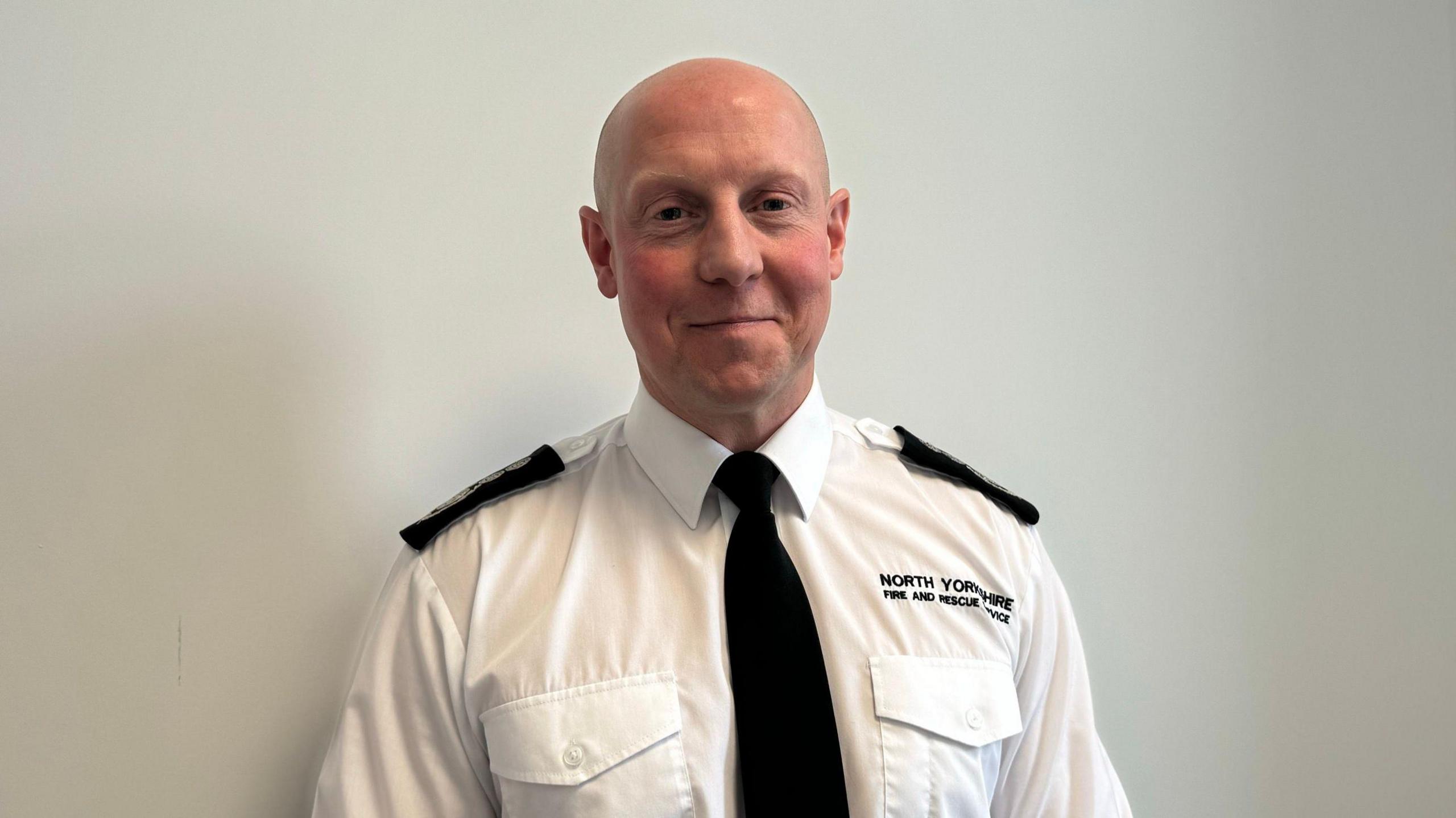 A man smiles at the camera wearing a North Yorkshire Fire and Rescue Service branded white shirt and black tie. He is clean shaven and has a bald head.