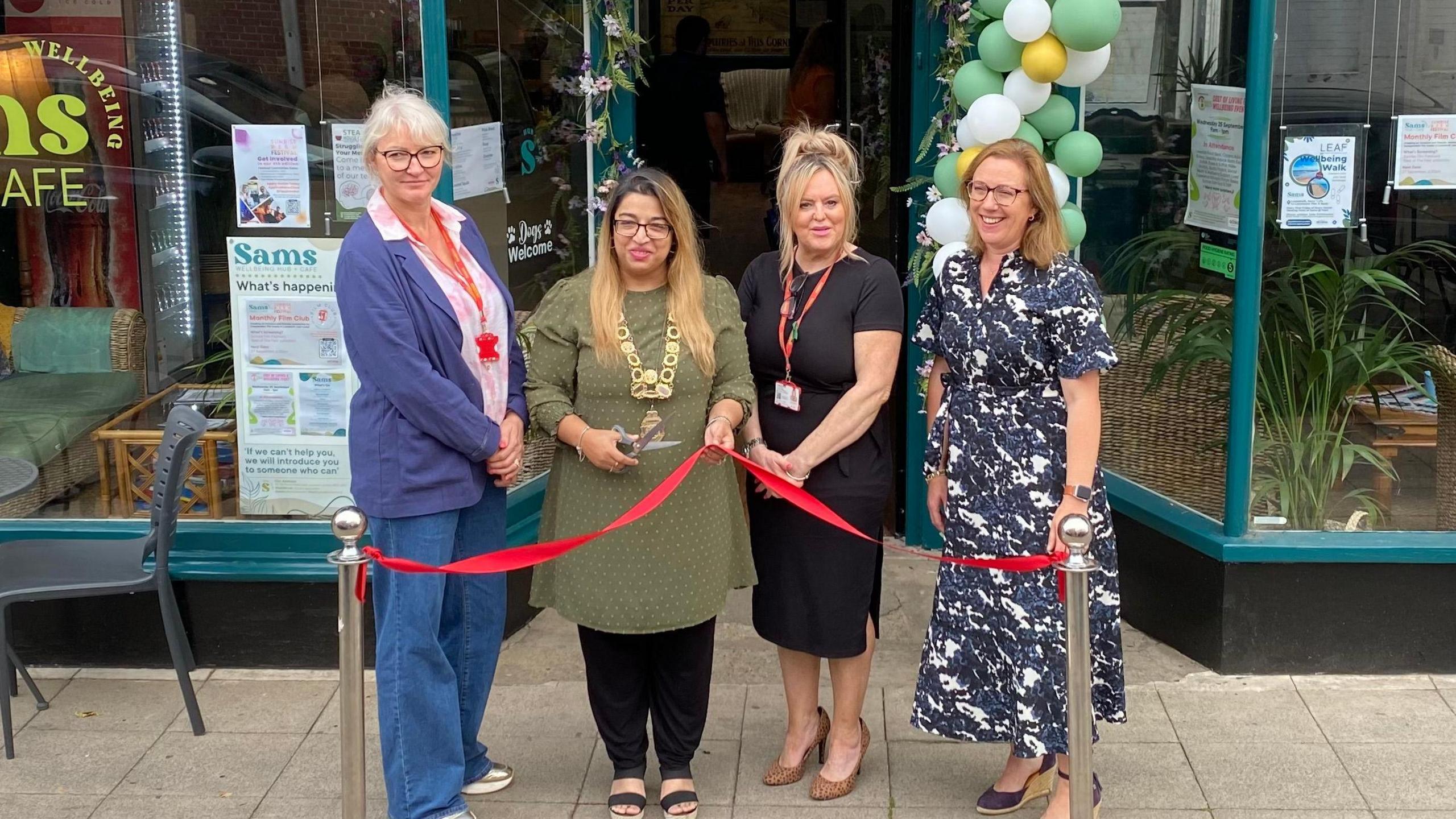 Four women are standing behind a red ribbon in front of the wellbeing centre which has large windows framed with green wood. A woman wearing a gold chain over a green dress and black trousers is holding a pair of scissors about to cut the ribbon.