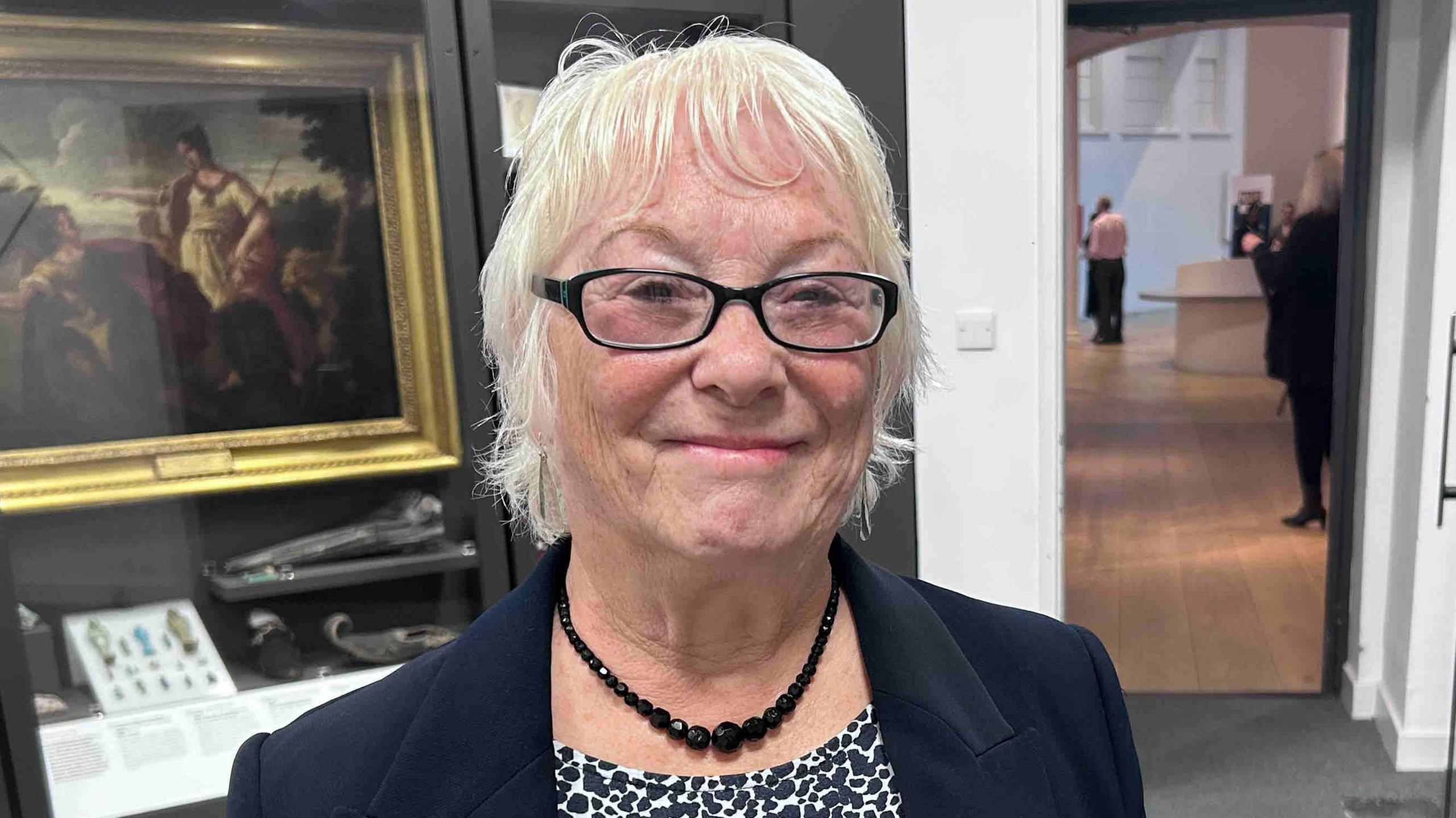 Danielle Stone is looking towards the camera smiling. She has short white hair and is wearing a black and white T-shirt, a black blazer and a black necklace. Behind her is a painting and a door leading to another room.