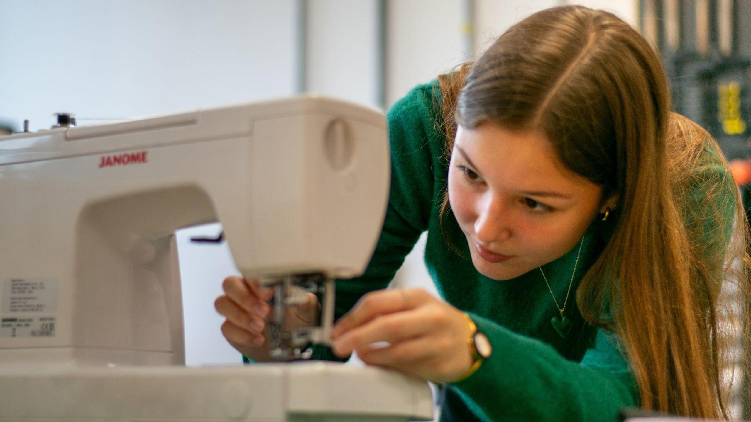 Former Great British Sewing Bee contestant Pascha Al-Qassab, wearing a green jumper, threads a sewing machine. She has long, light brown hair and is wearing a small watch.