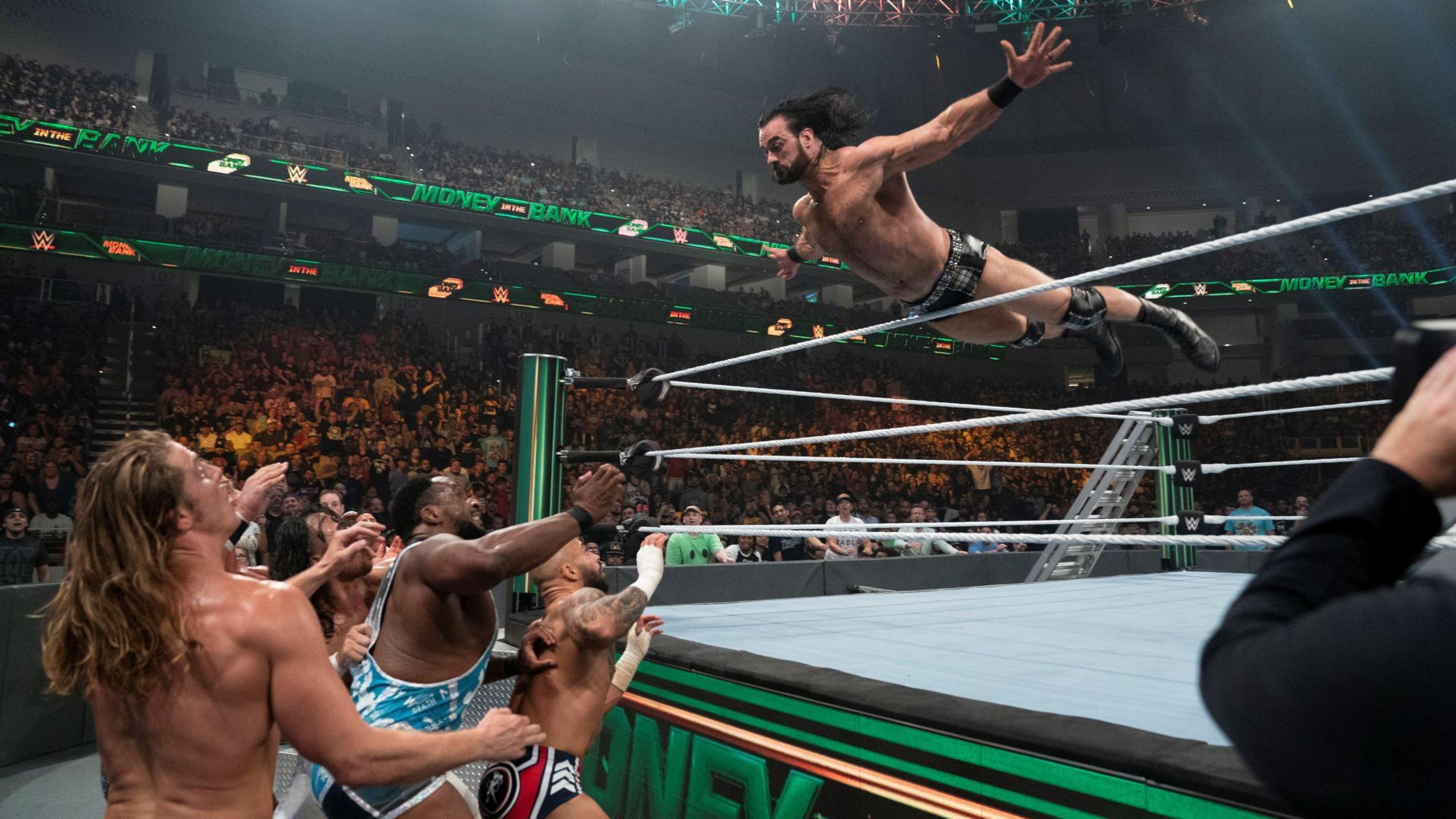 Drew McIntyre flies over the top rope, with several wrestlers waiting outside the ring for him to land on them. The ring apron and ropes have a green tinge to them, while a ladder is propped up against one of the other turnbuckles outside the ring.