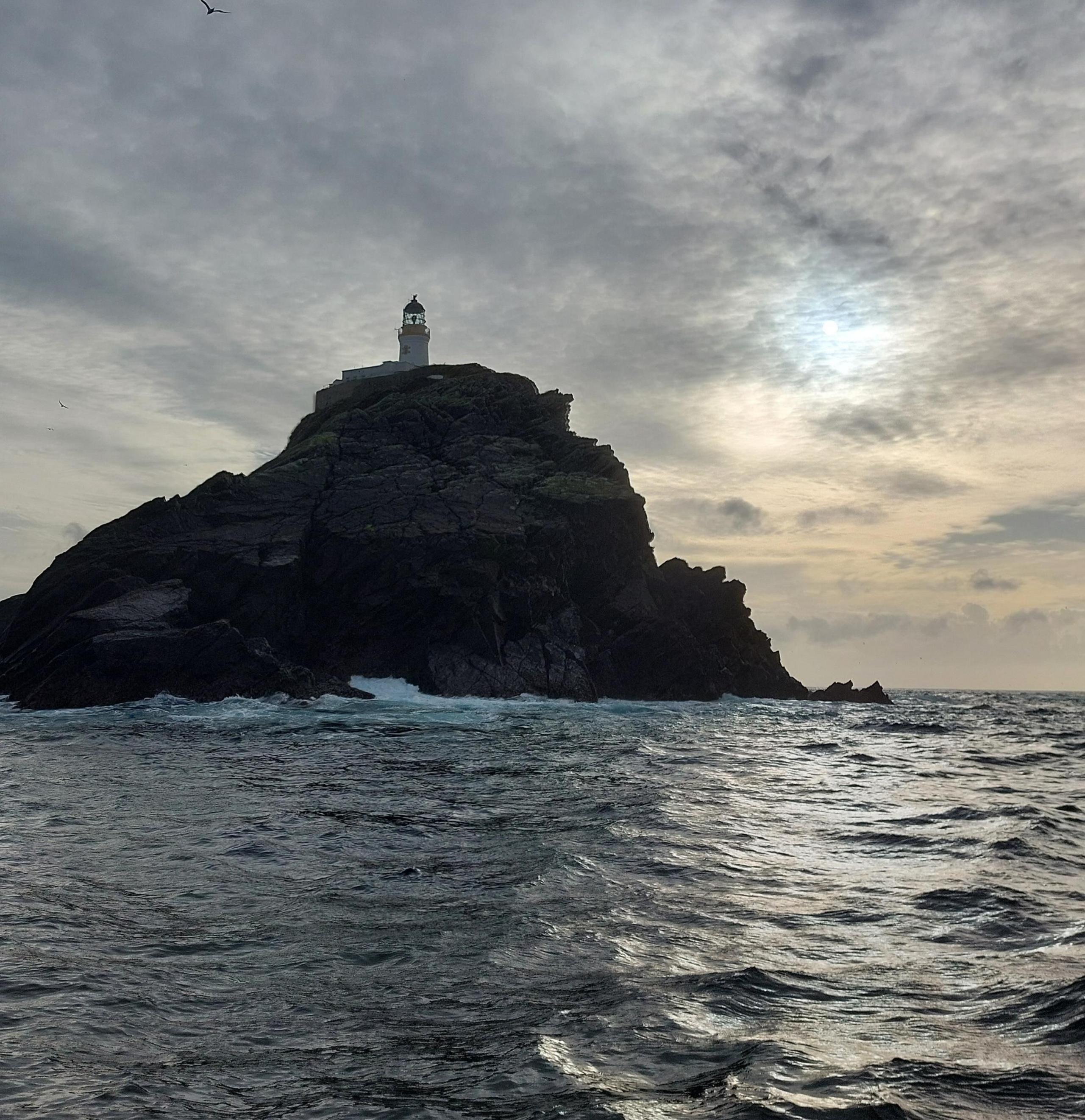 A lighthouse on a small, rocky island with choppy sea in the foreground. The sky has hues of orange.