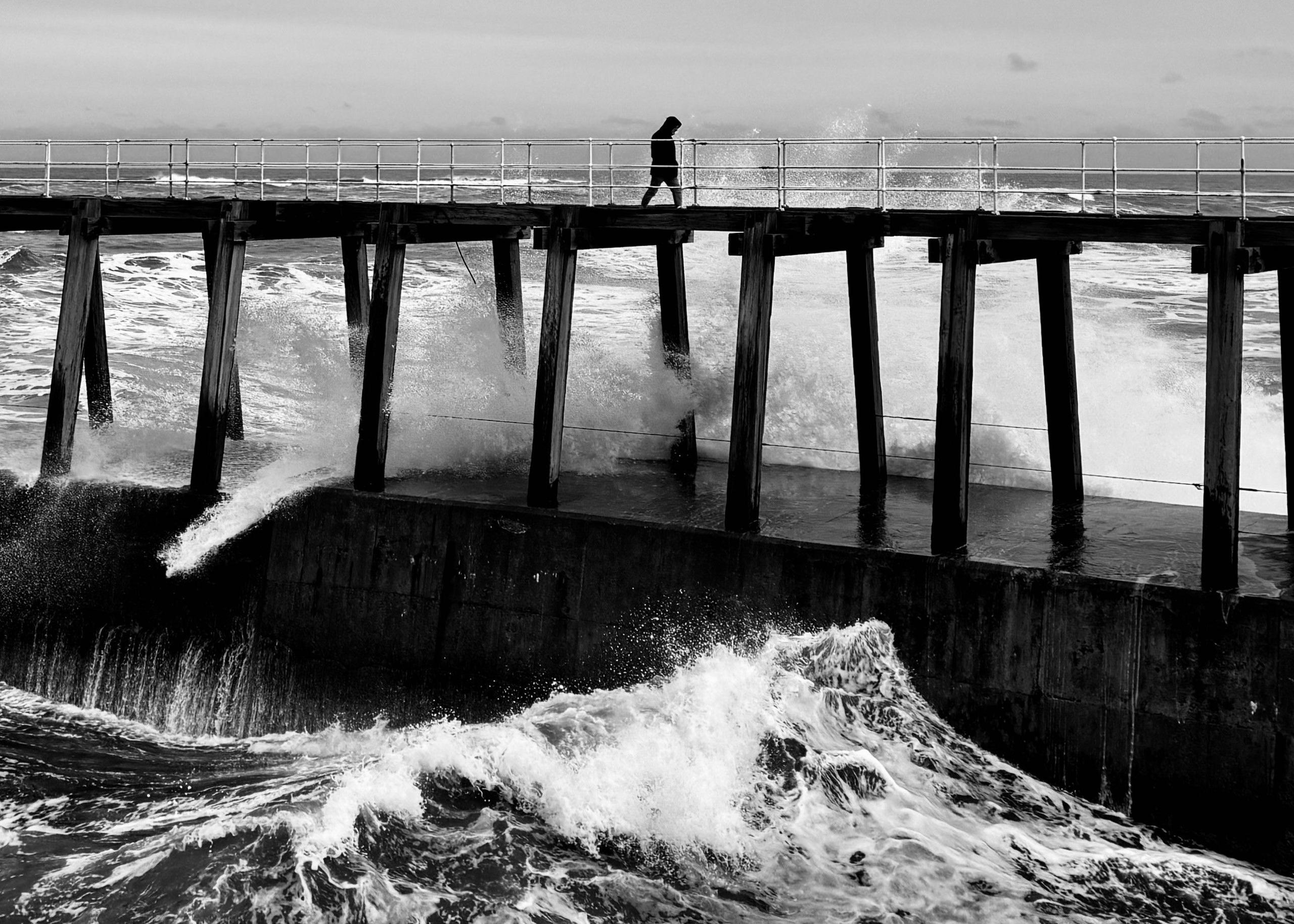 A figure walks along a pier over the sea as waves break below