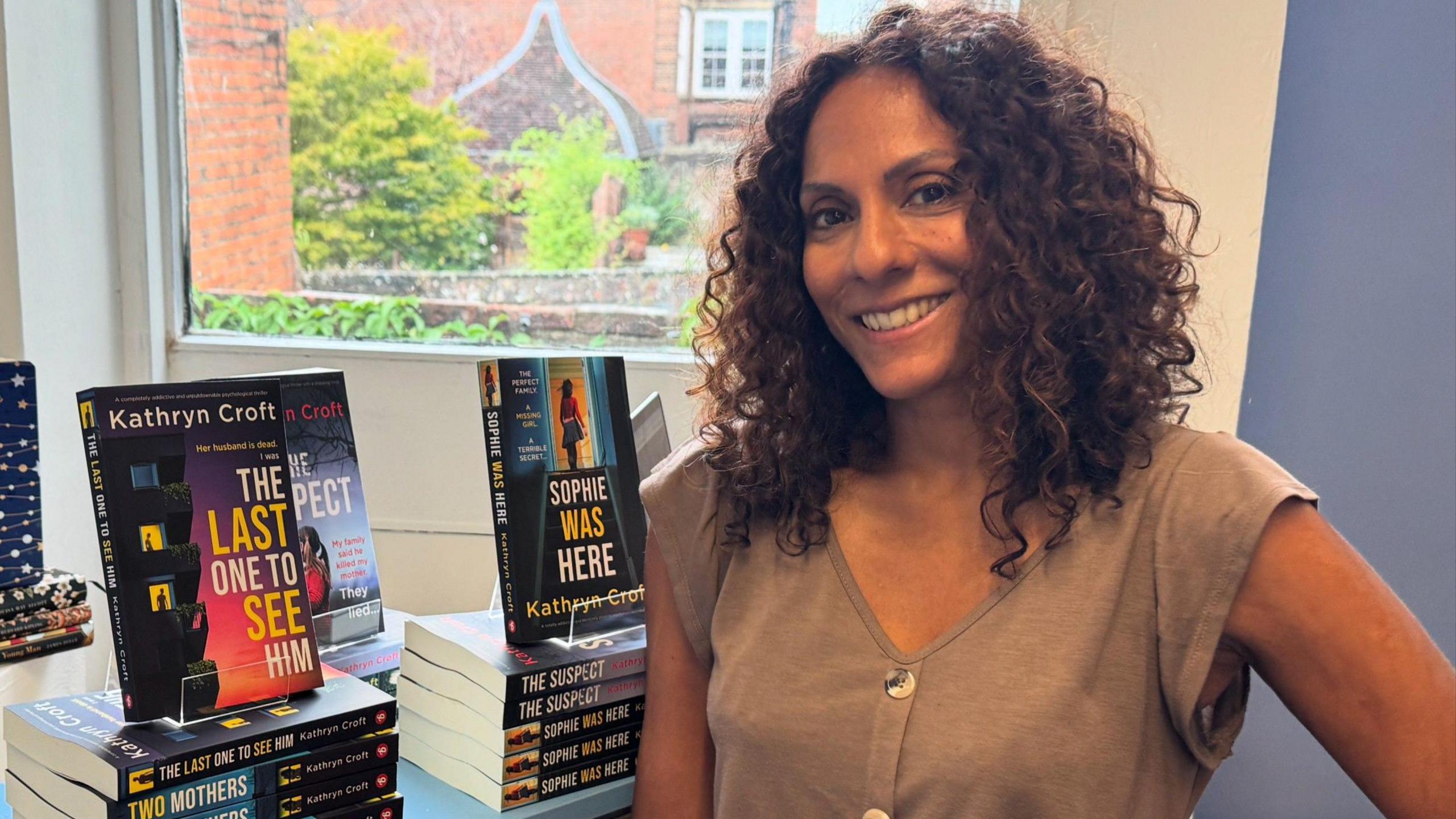 Kathryn Croft, a woman with long dark curly hair, wears a light brown sleeveless top as she stands in front of several hardback copies of her book "The Last One To See Him", in a bookshop publicity photograph.