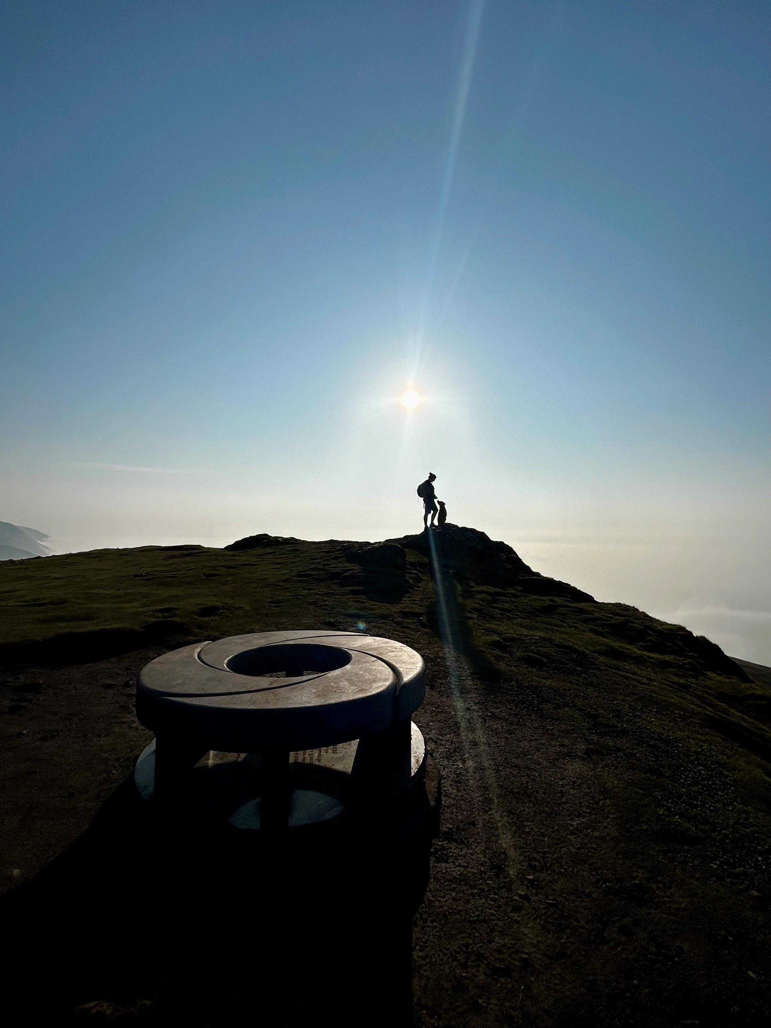 A silhouetted woman and dog on top of a hill with a circular monument in the foreground.