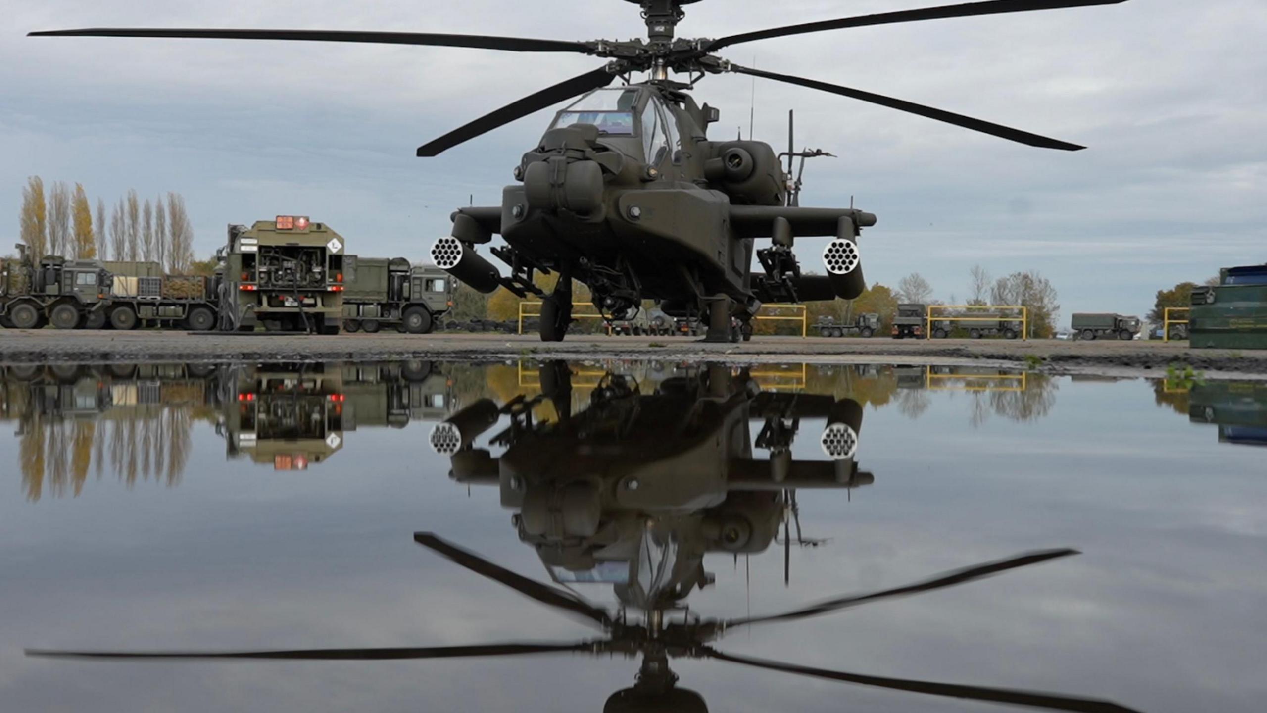 A helicopter sits on an airfield. Its reflection can be seen in a puddle of rainwater than sits in front of it. Other army vehicles are parked behind the helicopter.