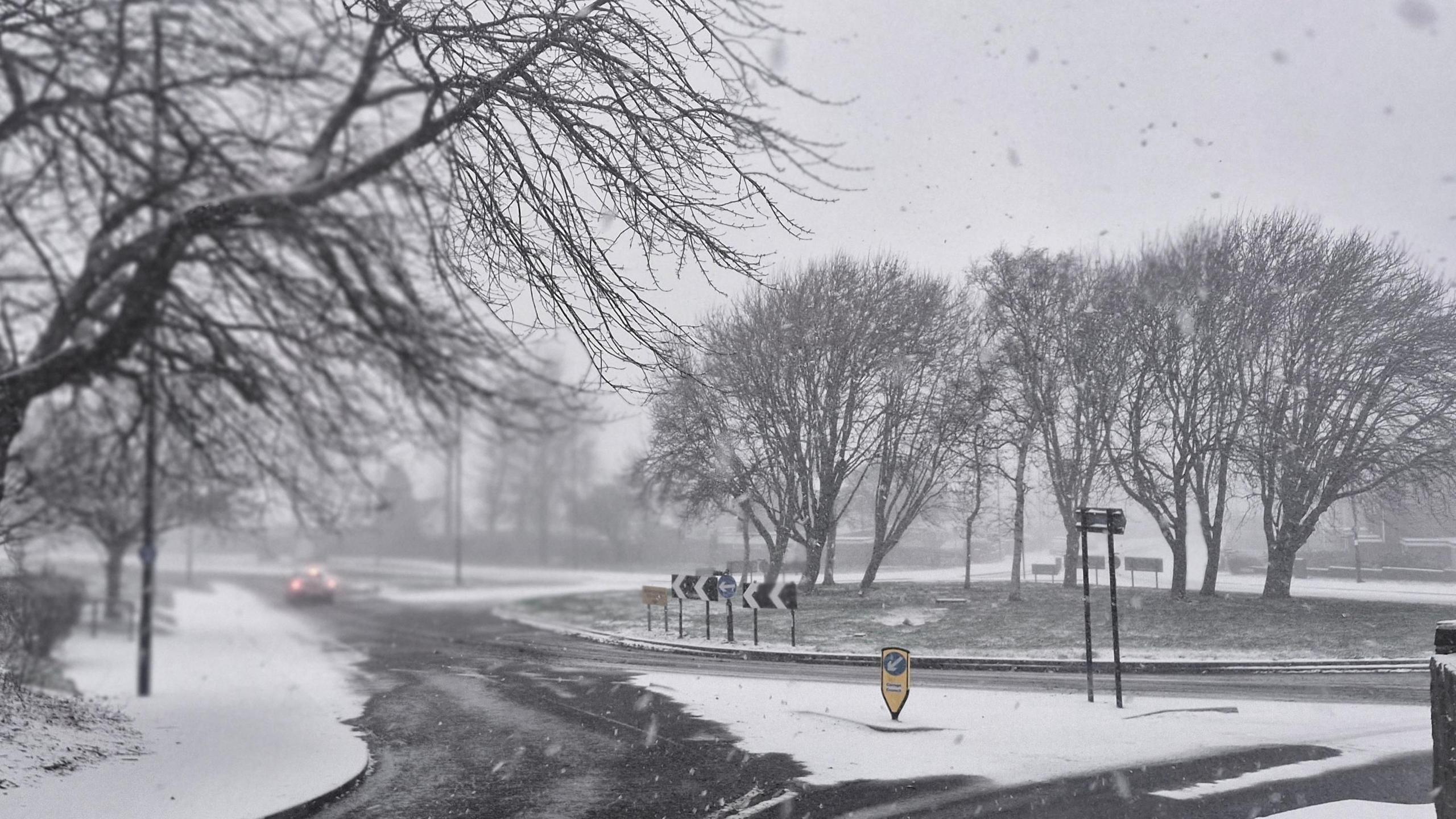 Slippery looking roads and snow-covered pavements surround a roundabout. Only one car visible in the distance.