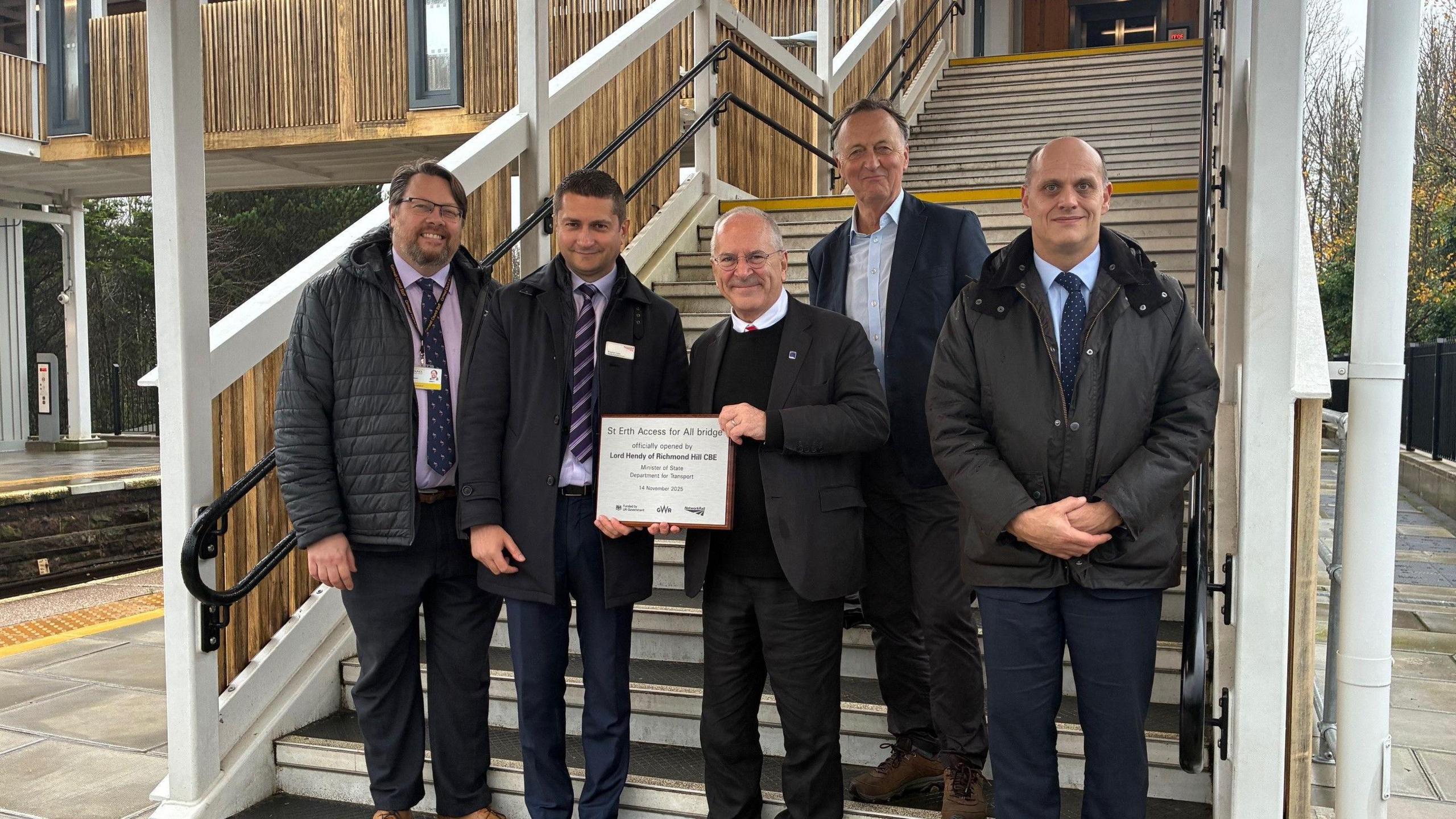 Five men in suits stand at the foot of the stairs of the new bridge. The transport minister Peter Hendy holds a plaque.