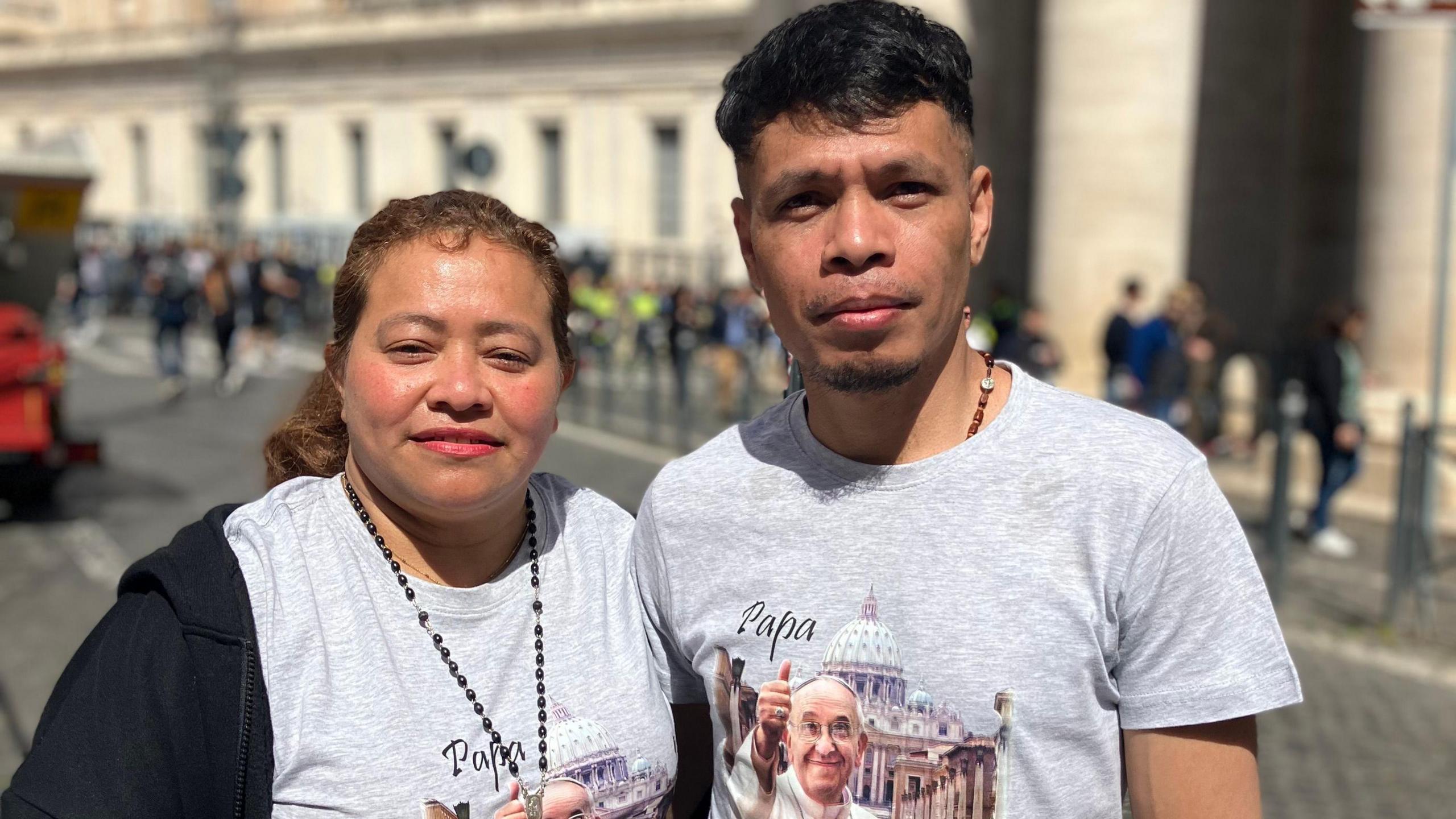 Crispino and Maria Carmo stand side by side, looking to the camera. They both wear grey T-shirts with a print of Pope Francis on them. in the print, the Pope is wearing a white gown and glasses while making a thumps-up gesture. The T-shirts say Papa on them. Crispino and Maria both have a beaded rosary chain round their neck.
