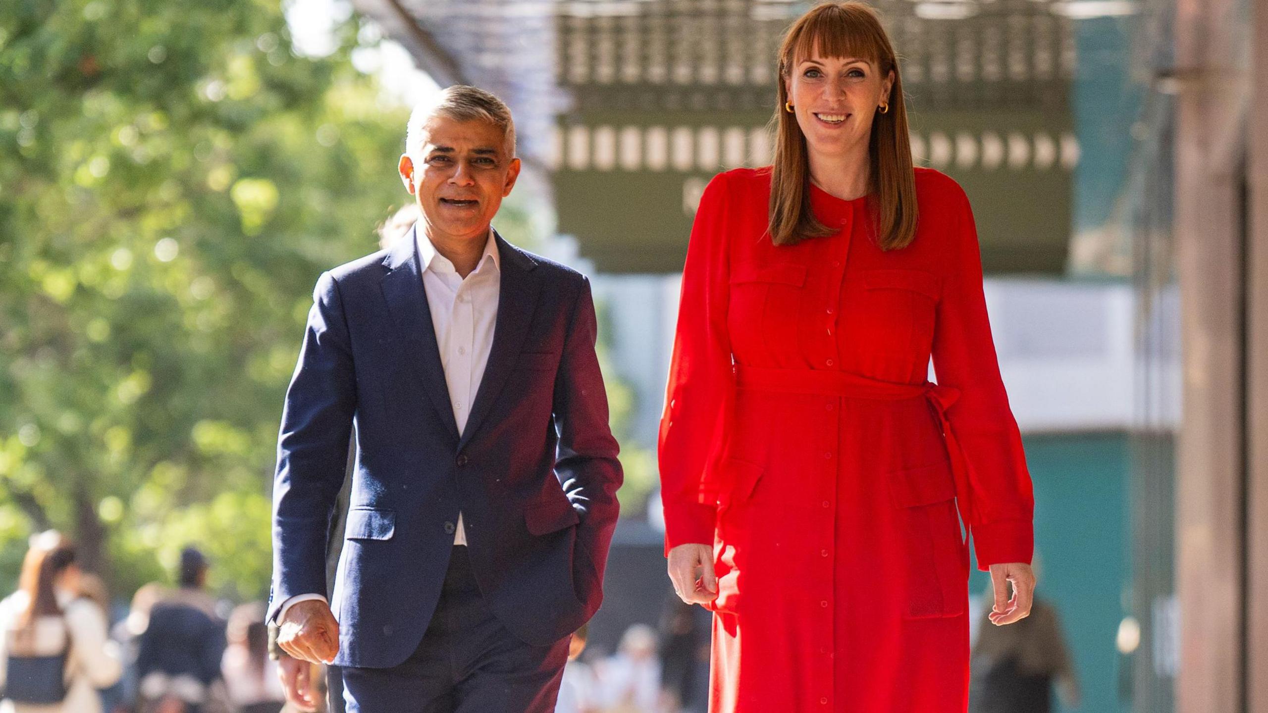 Sir Sadiq Khan, in a dark blue suit and white shirt and Angela Rayner in a bright red dress walk side by side in the sunshine down a London street. They are both smiling. 