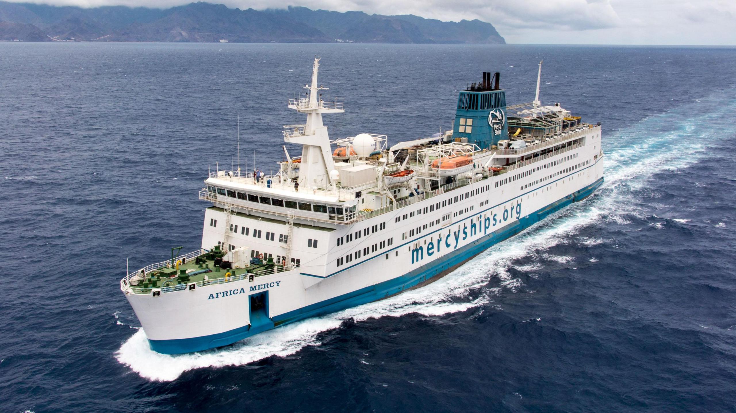 A photo which shows the ship the Africa Mercy, The ship has a blue stripe at the bottom of it, and the rest is white. On the side in blue is written mercyships.org and Africa Mercy on the front.