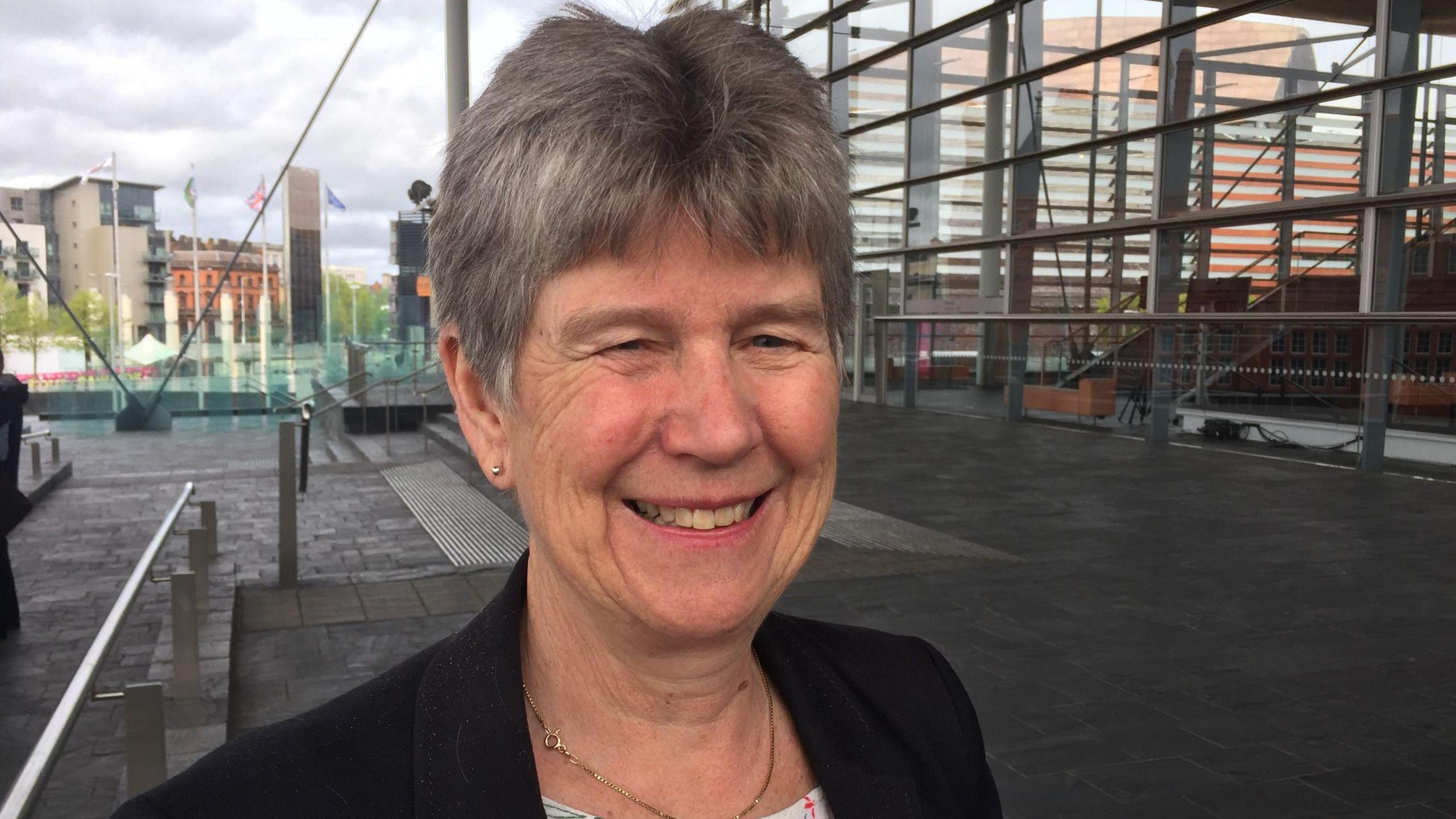 Jane Hutt photographed outside the Senedd, she has short silver hair, blue eyes and smiles towards the camera. She is wearing a black blazer and gold stud earrings and a gold slim chain necklace. 