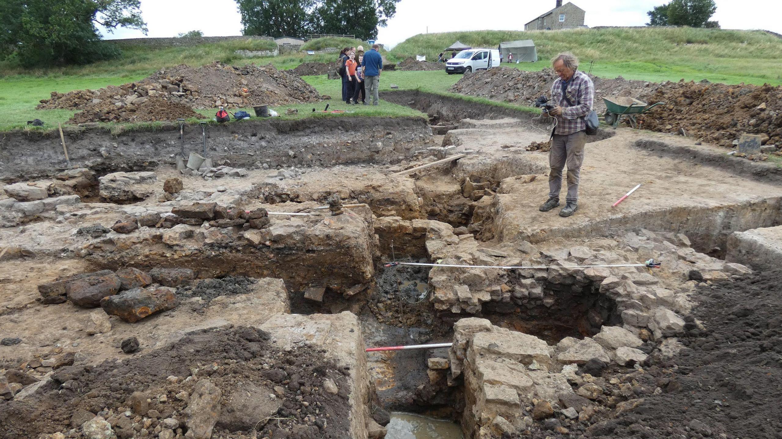 The picture shows the site of a dig. The ground has been dug up and there are stones that appear to be the inside of a fort. People are standing on top and there are piles of mud next to them. One man is taking a photo. In the background is the countryside.