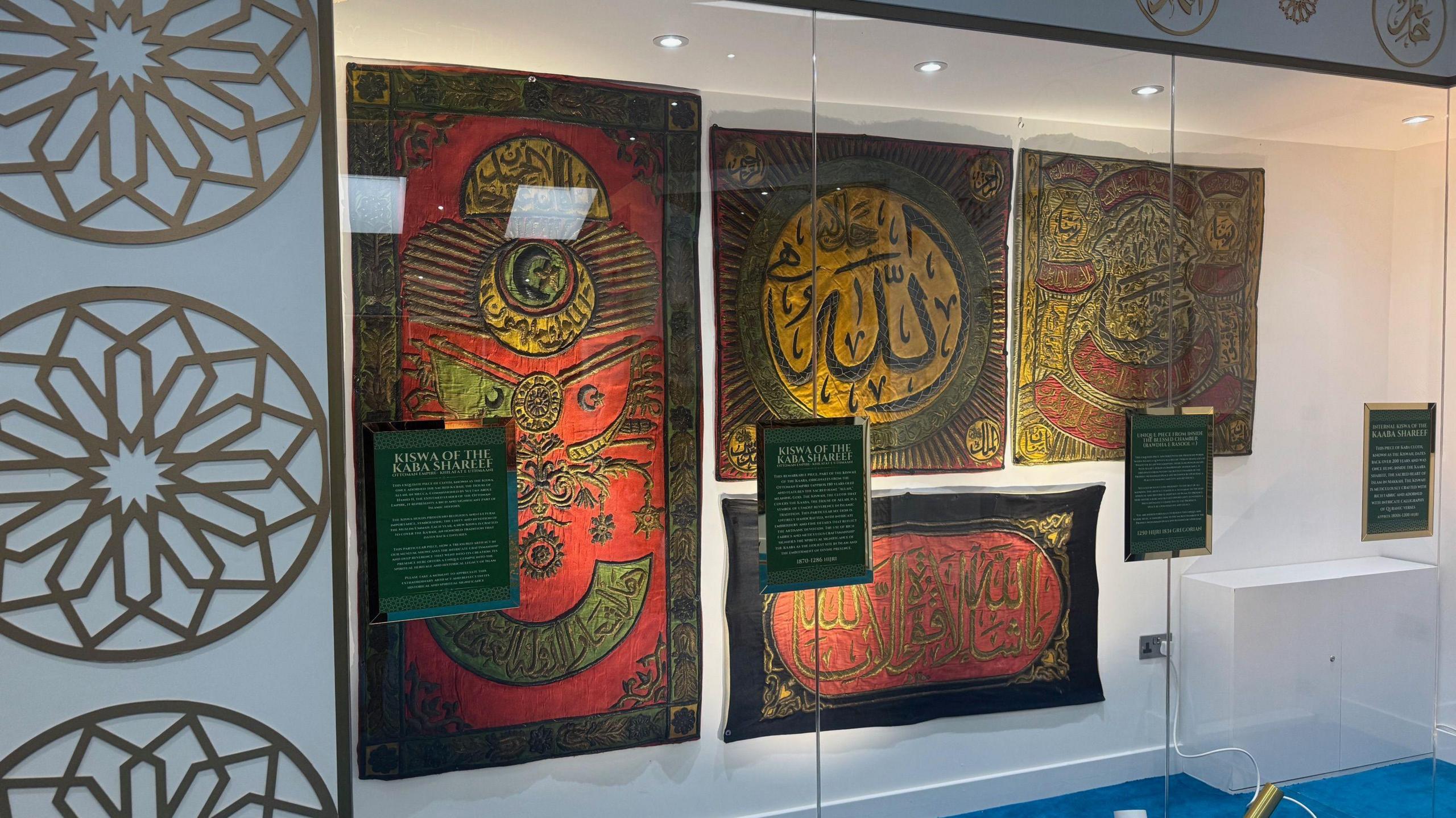 A glass cabinet, lit, features four colourful prayer mats 