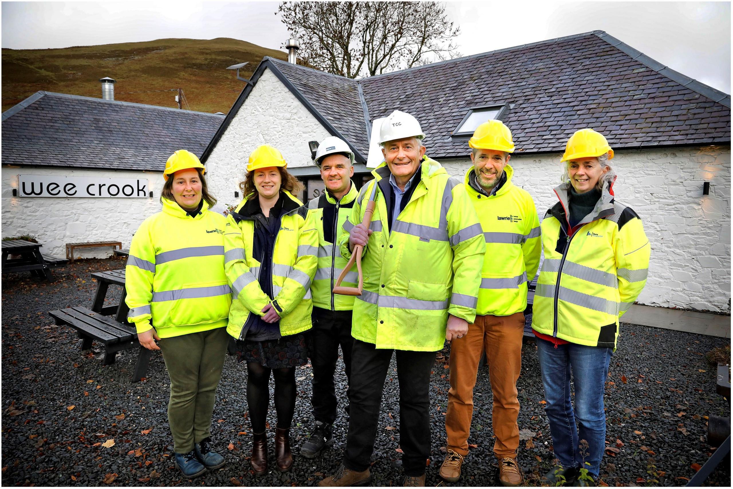 A line-up of six people in high visibility gear, one of them carrying a shovel, outside the Wee Crook cafe - a white building with a black slate roof
