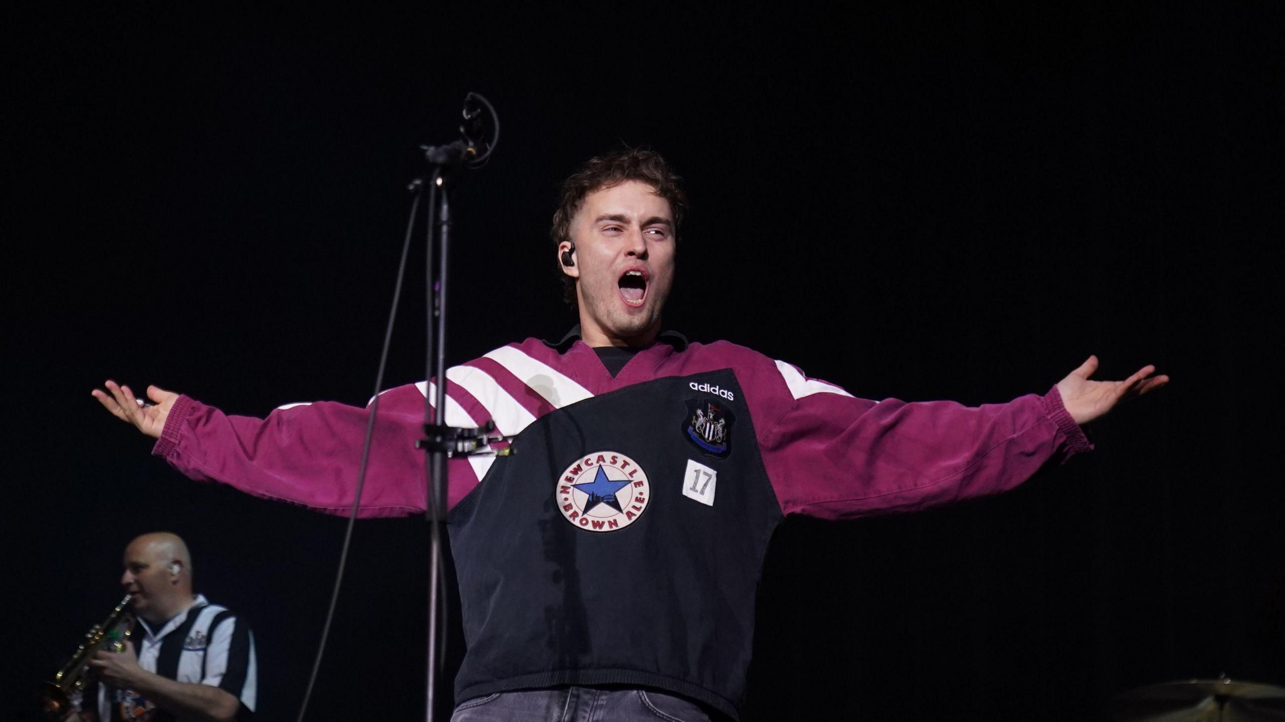 Sam Fender on stage. He is wearing a Newcastle United top which contains the club logo as well as the Newcastle Brown Ale star. A saxophone player in the background is wearing a Newcastle United shirt.