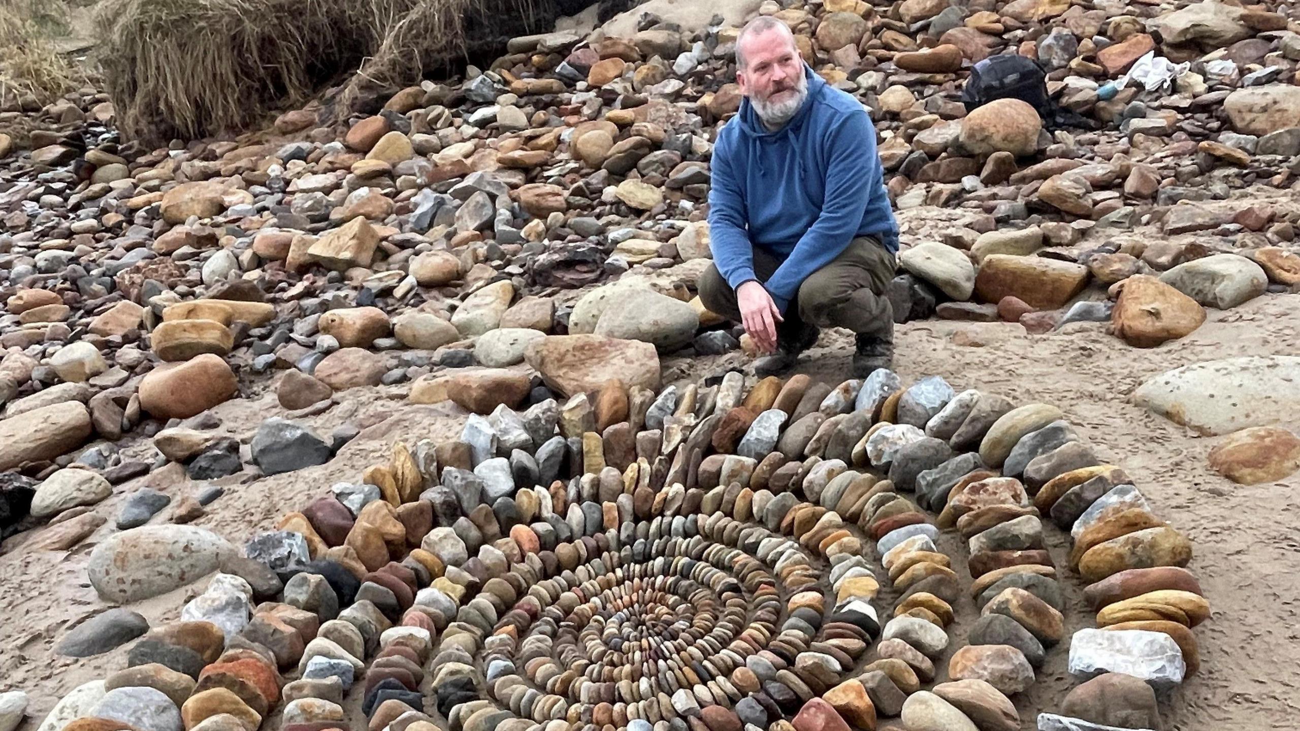 The artist James Brunt is sitting on his haunches in front of his artwork which is a large circle of stones beginning with very small ones and stretching out to create a large structure. He is a man in his 50s with a grey beard and wearing a blue jumper  