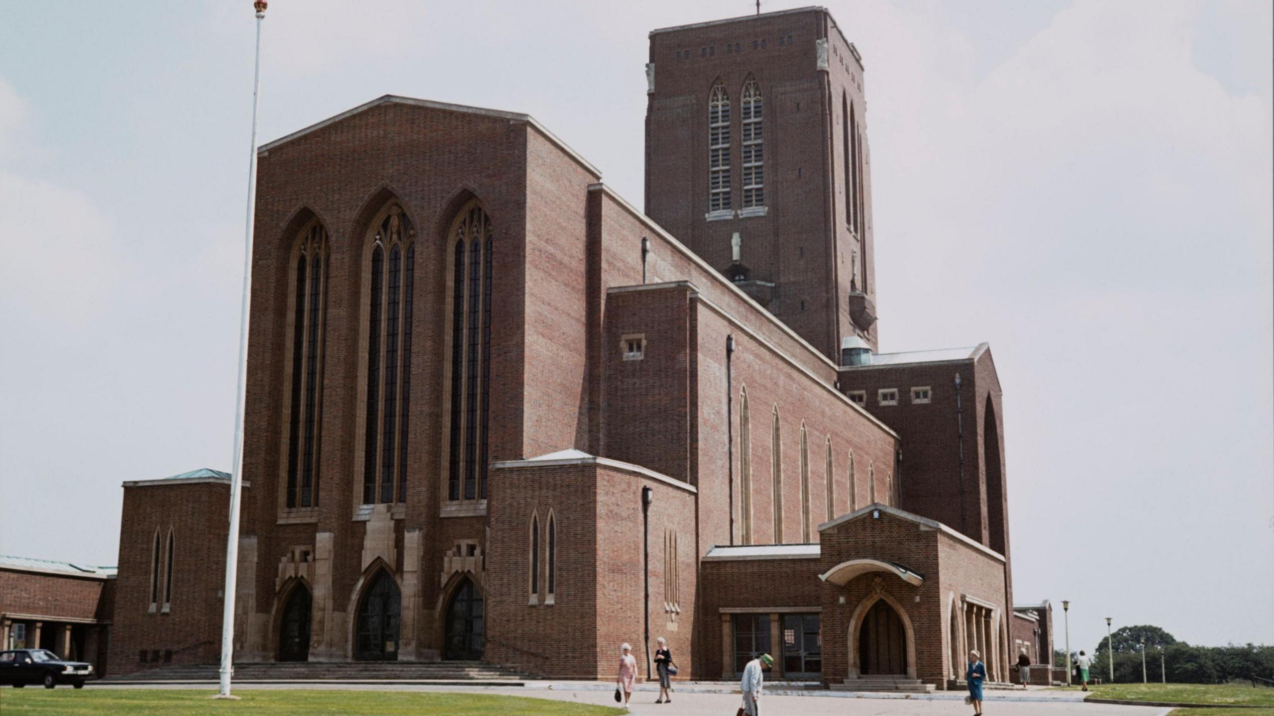 An austere brown brick cathedral with tall arched windows and sections of several different heights, including a tower. There is an area of green grass with a flag pole in front of the cathedral and a black car parked to the left. Six older people can be seen walking in the foreground. The sky looks murky and overcast, a muted blue/grey with big clouds.