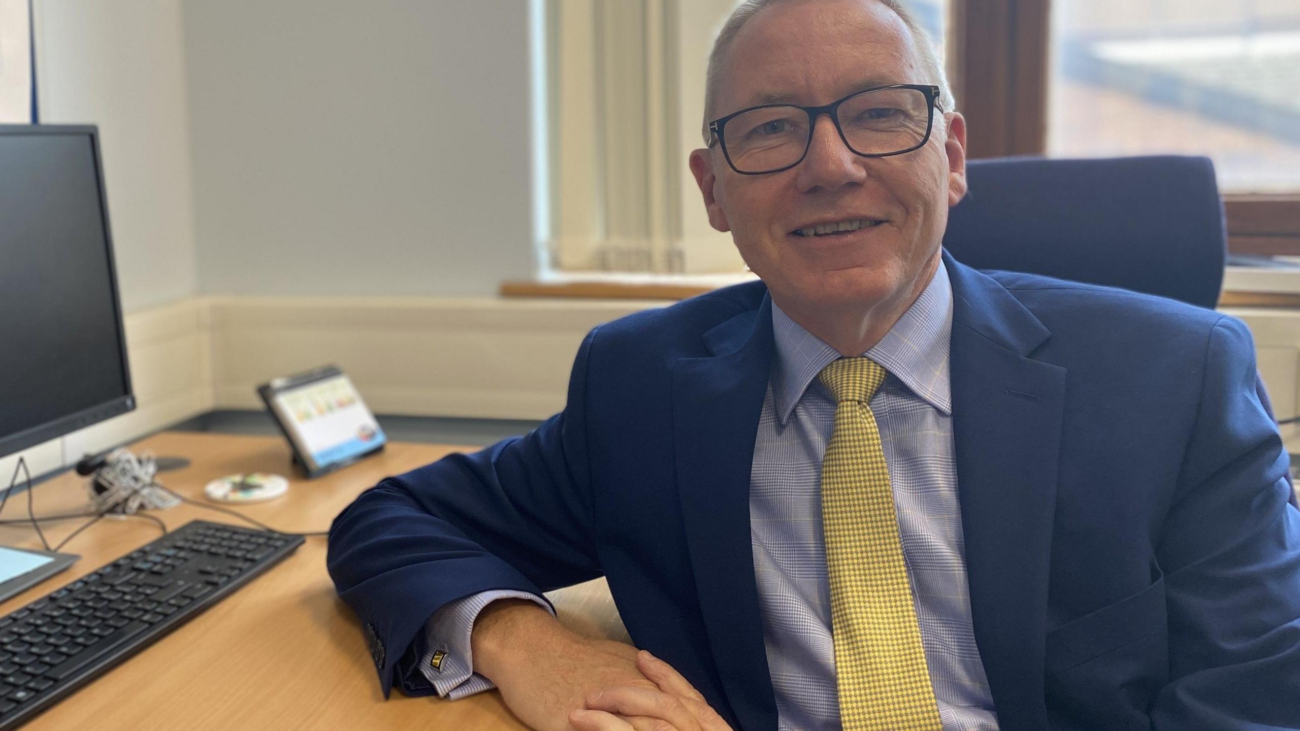 A man wearing a dark blue suit with a light blue shirt and a gold tie sits leaning on a desk whilst smiling for the camera