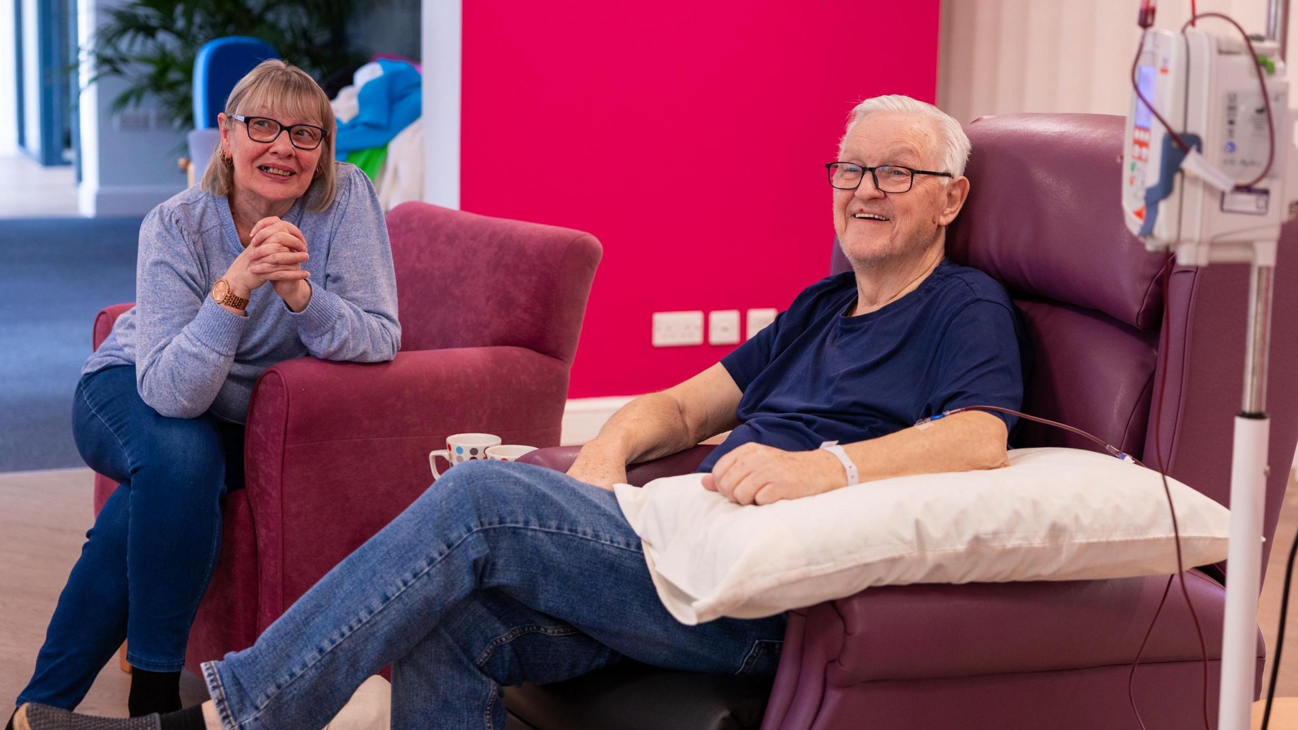 A man with white hair and wearing black glasses sits in a large purple padded chair, in what looks like a communal area of the hospice. A tube from his arm connects him to a portable medical machine with buttons and cables. He is looking at someone off camera and grinning. To his left a woman sits in another comfy chair leaning towards the man and looking at the person off camera. They both wear jeans and blue tops.