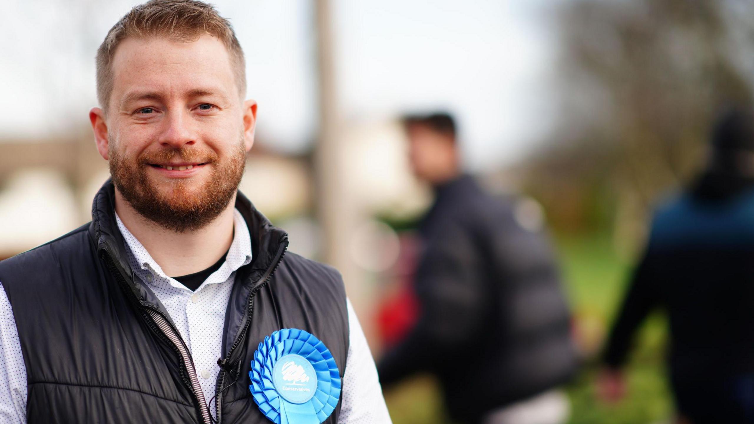 Sam Bromiley standing in a field wearing a black gilet and white shirt and a blue Conservative badge.