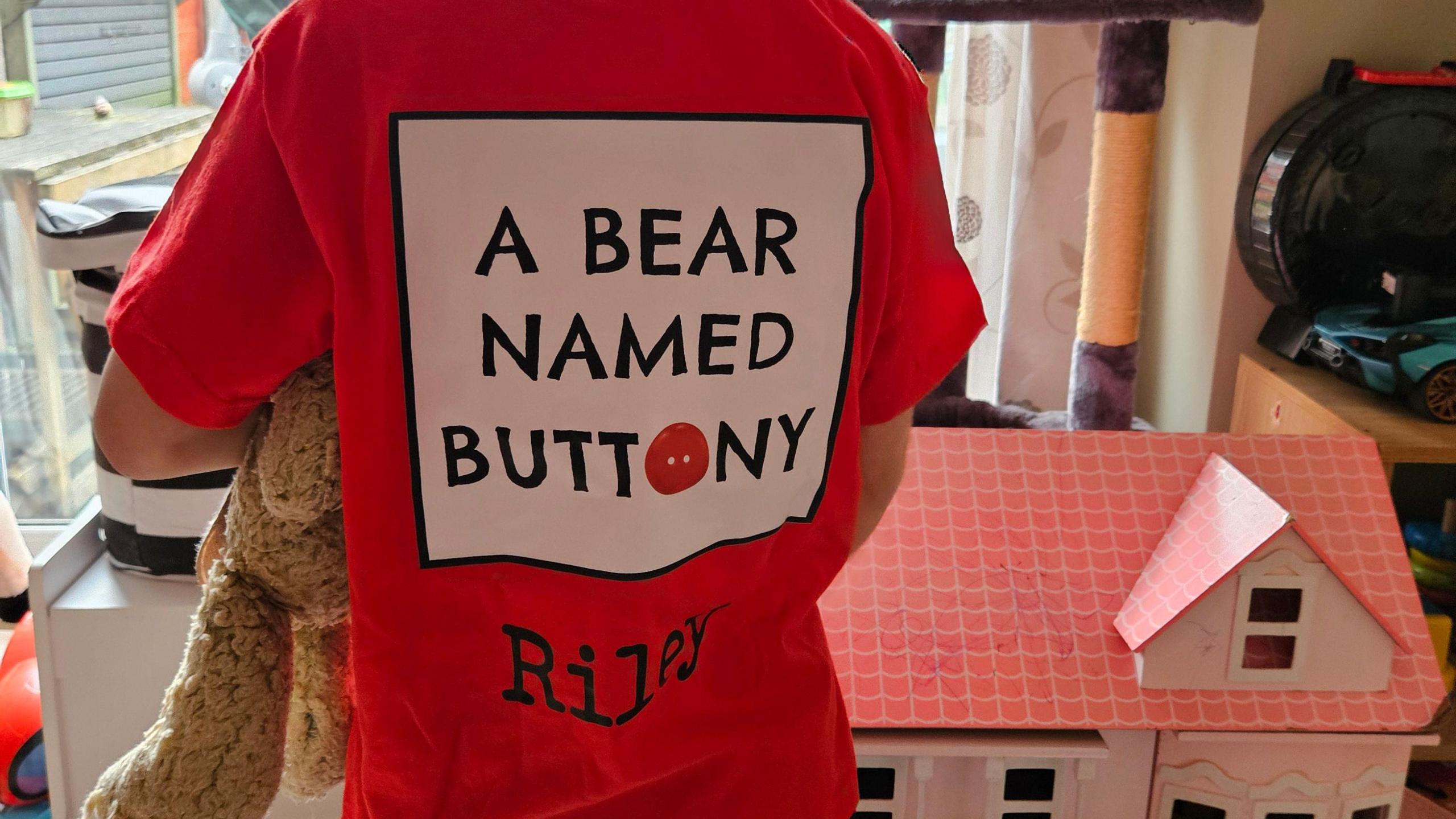 The back of Riley's red t-shirt which reads 'A Bear Named Buttony - Riley'. He is holding Buttony to his chest.