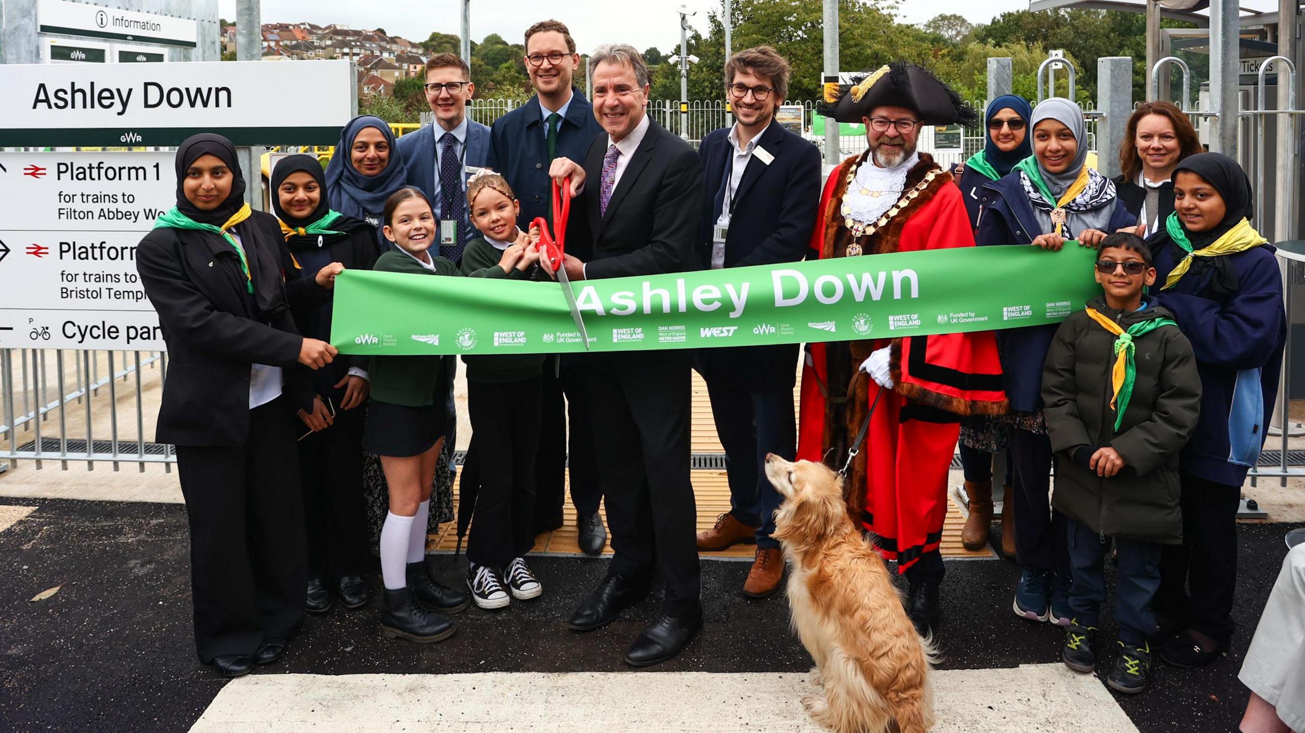 Various dignitaries, including the mayor of bristol, the mayor of the west of england Dan Norris and a group of school children holding a pair of giant scissors in front of a banner with the words "Ashley Down", preparing to cut the banner