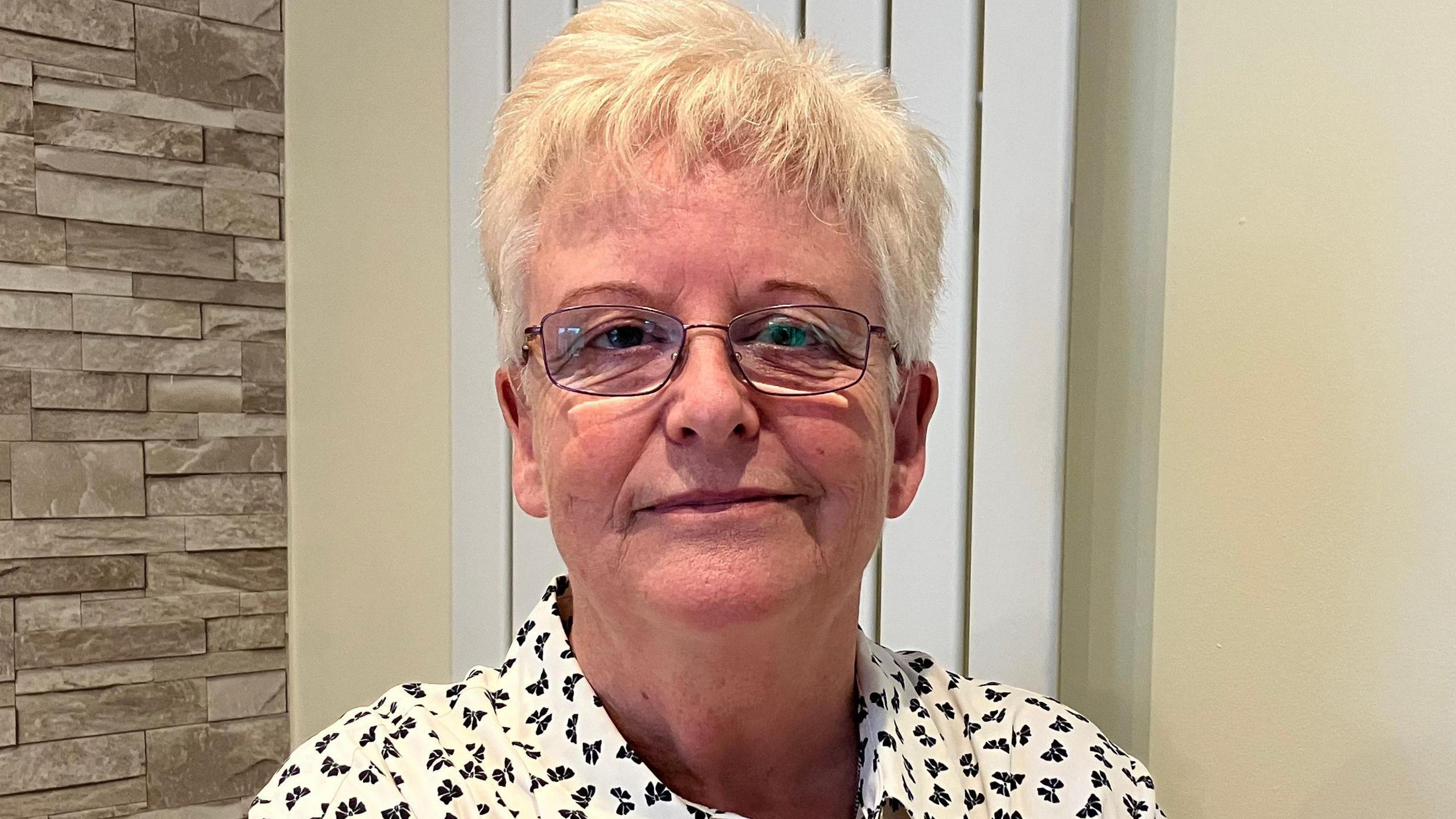 Maggie Lockhart seated at a table at home. She is wearing glasses and a white shirt with a black pattern. She is sitting on a wooden chair, and another chair is partially visible in the bottom right corner of the image.
The background features two distinct wall textures: on the left, there is a wall with a stone-like appearance, while on the right, the wall is light-coloured and has a framed picture hanging on it. The framed artwork depicts a scenic landscape with trees, water, and possibly some buildings in the distance.