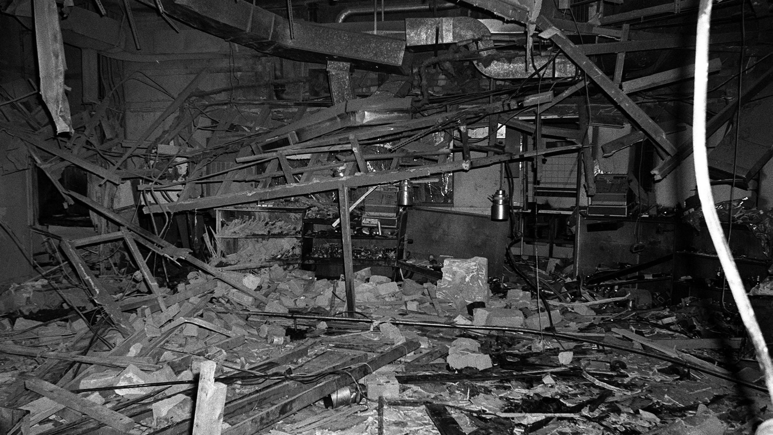 A black and white image of the inside of one of the pubs. There is rubble everywhere, with structural metal parts of the building having fallen down, windows are smashed. There are pipes, boards, glass and wires strewn over the floor.