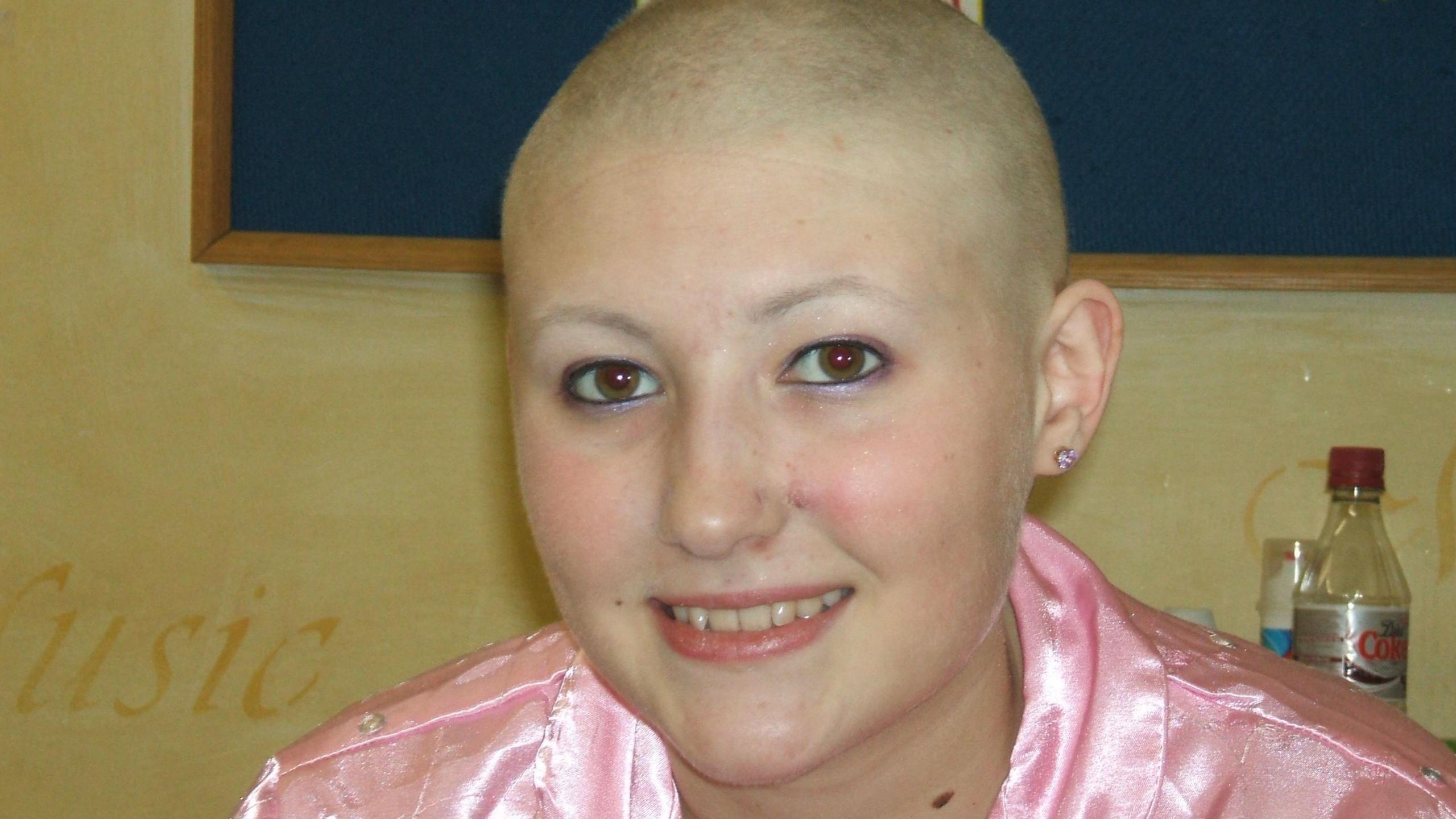 A 14-year-old girl with a shaved head, she is wearing a shiny pink jacket, behind her an empty bottle of diet Coke is visible