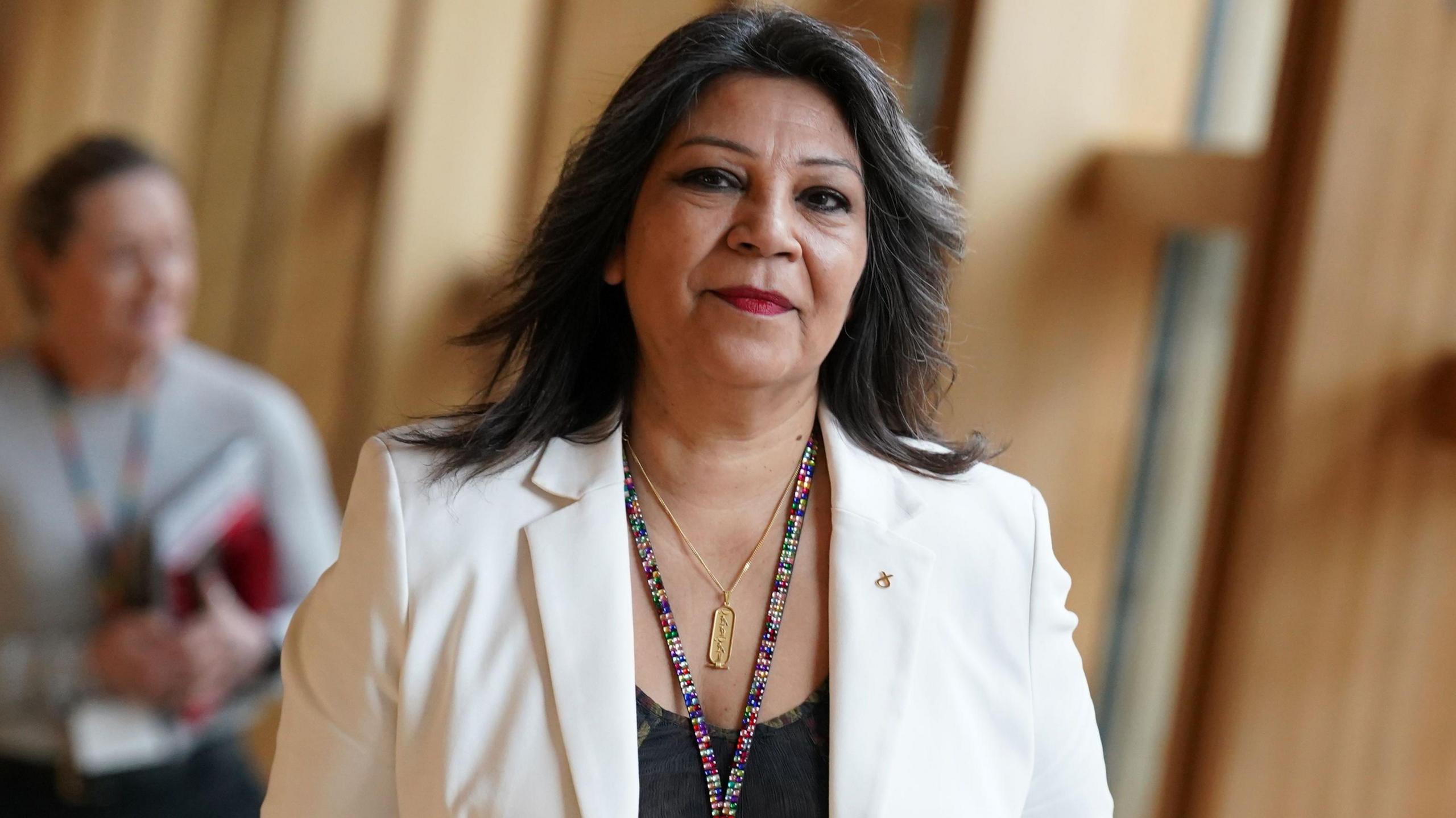 Equalities Minister Kaukab Stewart walking through the wood panelled foyer of the Scottish Parliament building in Edinburgh. She is looking directly att he camera and a man in blurred in the background. She is wearing a white blazer-style jacket, black top and gold chain and pendant. She has long dark hair, with specks of grey, and is looking directly at the camera.