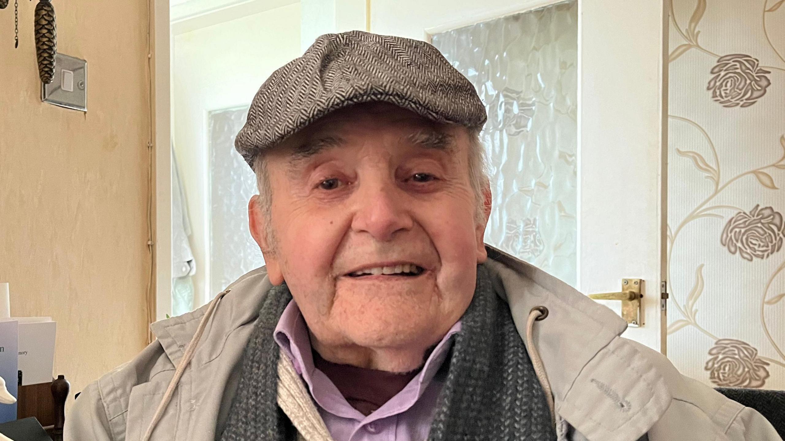 John looking down the camera, he's wearing a tweed flat cap and a beige overcoat, with a jumper, shirt and scarf underneath. He's sat in his front room, you can see patterned wallpaper and the hallway door behind him. 