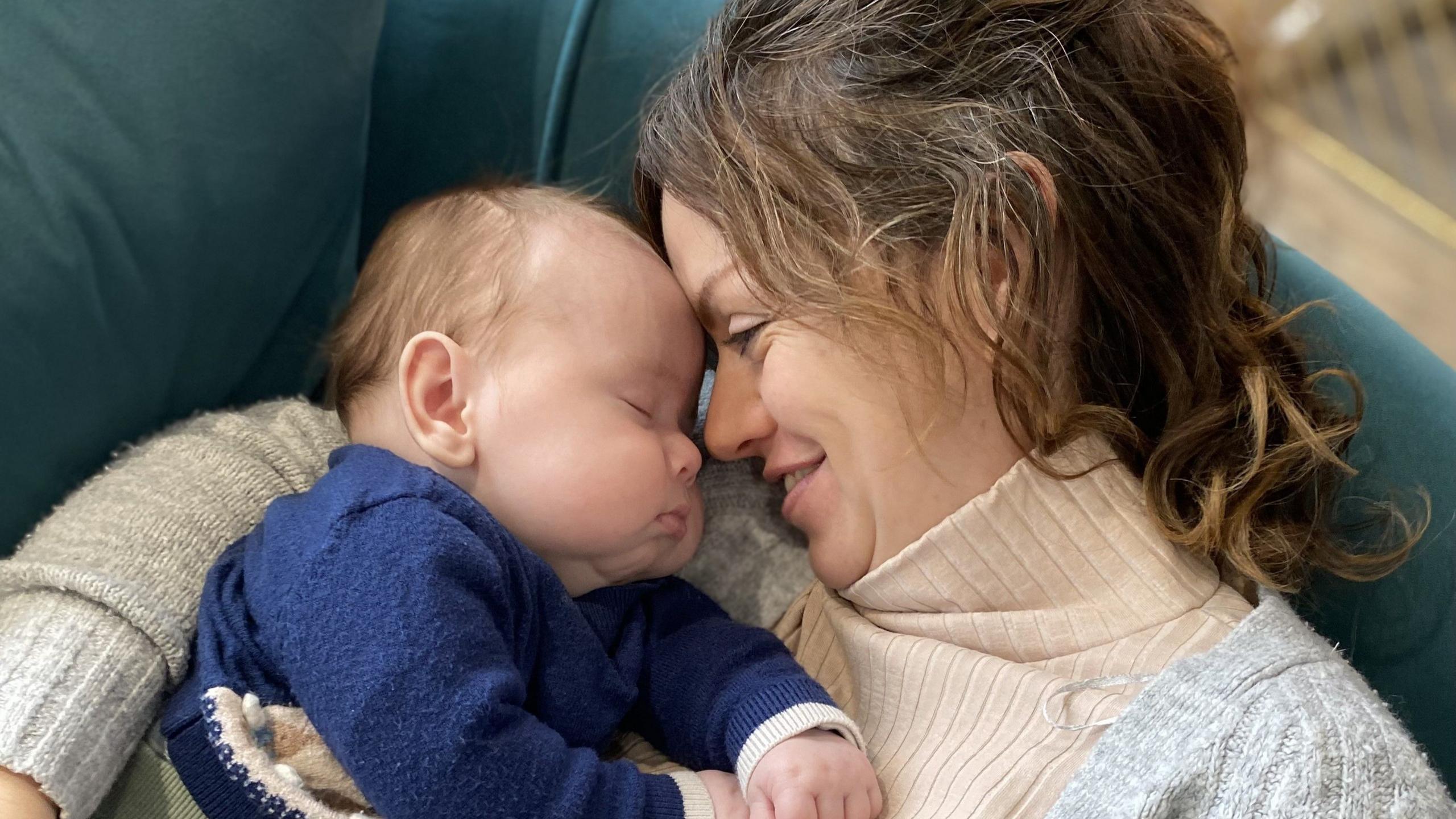 Image of a woman cuddling a sleeping baby. She has her hair tied back, is wearing a roll neck jumper and is smiling. She has her faced pressed against the baby's, which has its eyes closed. The baby is wearing a dark coloured jumper and has very chubby cheeks.