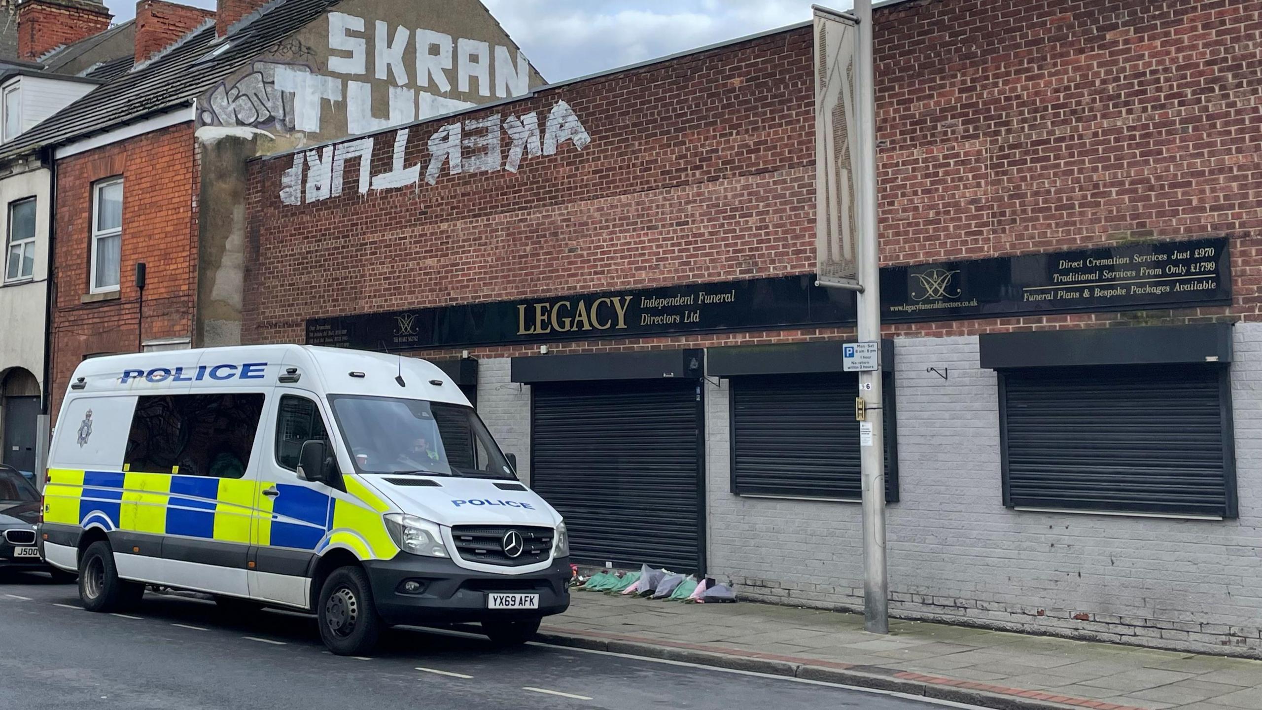 A white police van, with yellow and blue decor, outside the premises of Legacy Independent Funeral Directors, in Hessle Road, Hull. The large, red-brick building has black shutters, a flat roof and a black and gold "Legacy" sign. White graffiti covers part of the front wall, with more graffiti on a neighbouring roof. 