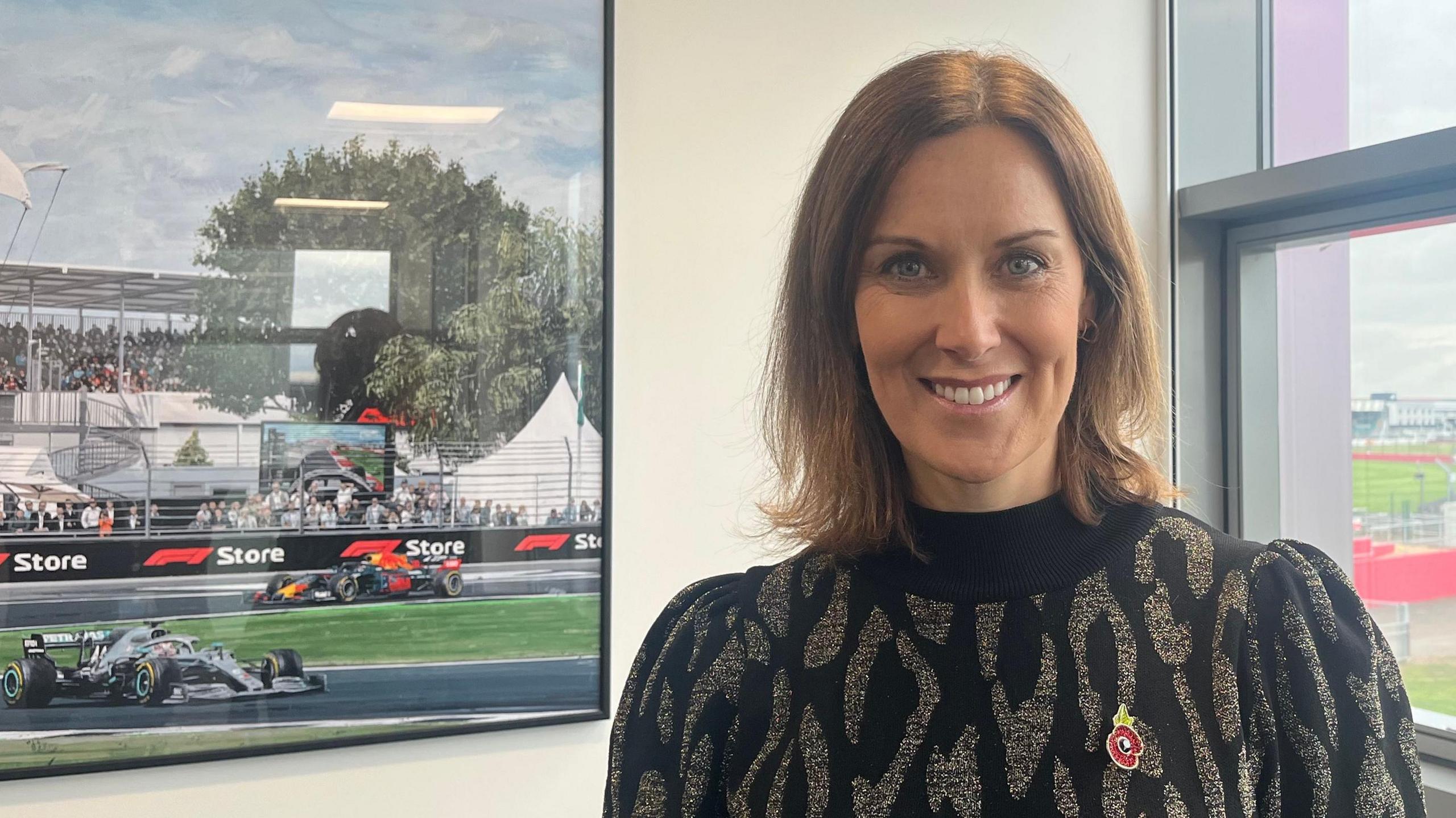 Lee McKenzie is smiling at the camera in front of a framed photograph of the racing track. She has brown hair and is wearing a leopard print jumper with a poppy badge for Remembrance Day. 