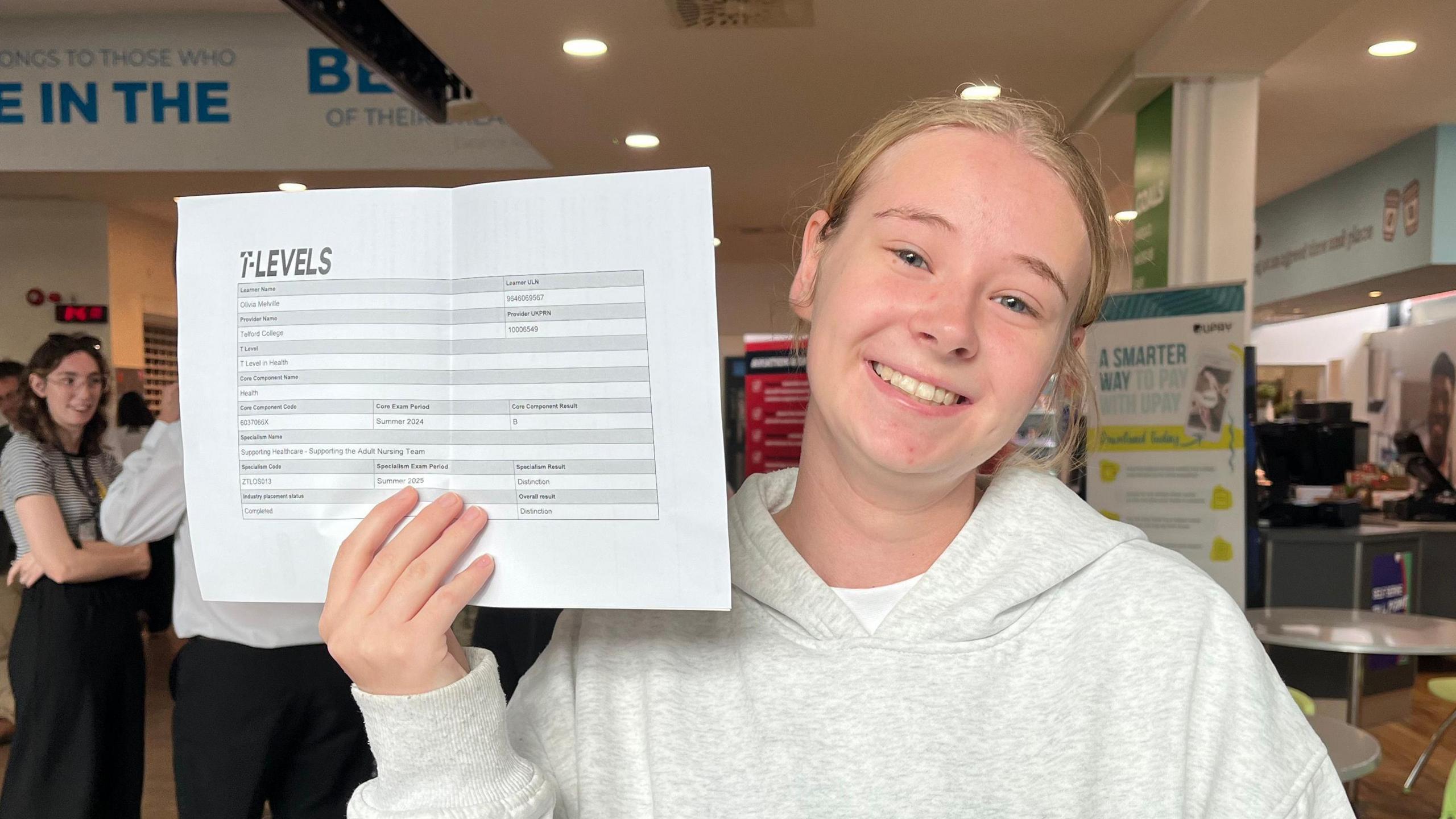 Olivia looking into the camera and smiling. She's wearing a grey hoodie and is holding up a landscape piece of paper - you can't make out what's written on it but at the top, in bold, is written 'T Levels.'