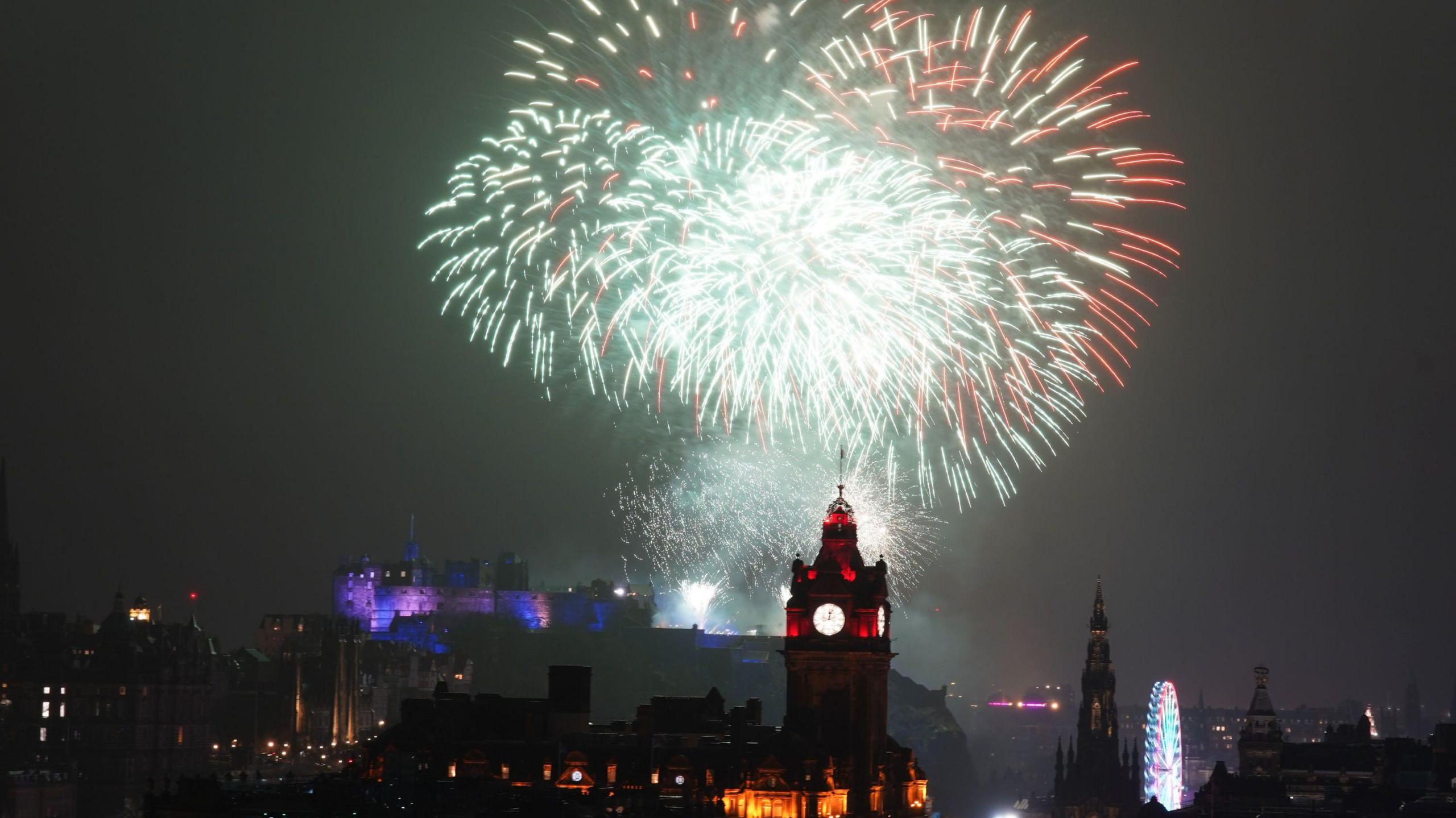 Fireworks, which are green and red, explode over the Balmoral Hotel in Edinburgh. The castle is in the background and is lit up in purple. The sky is dark.