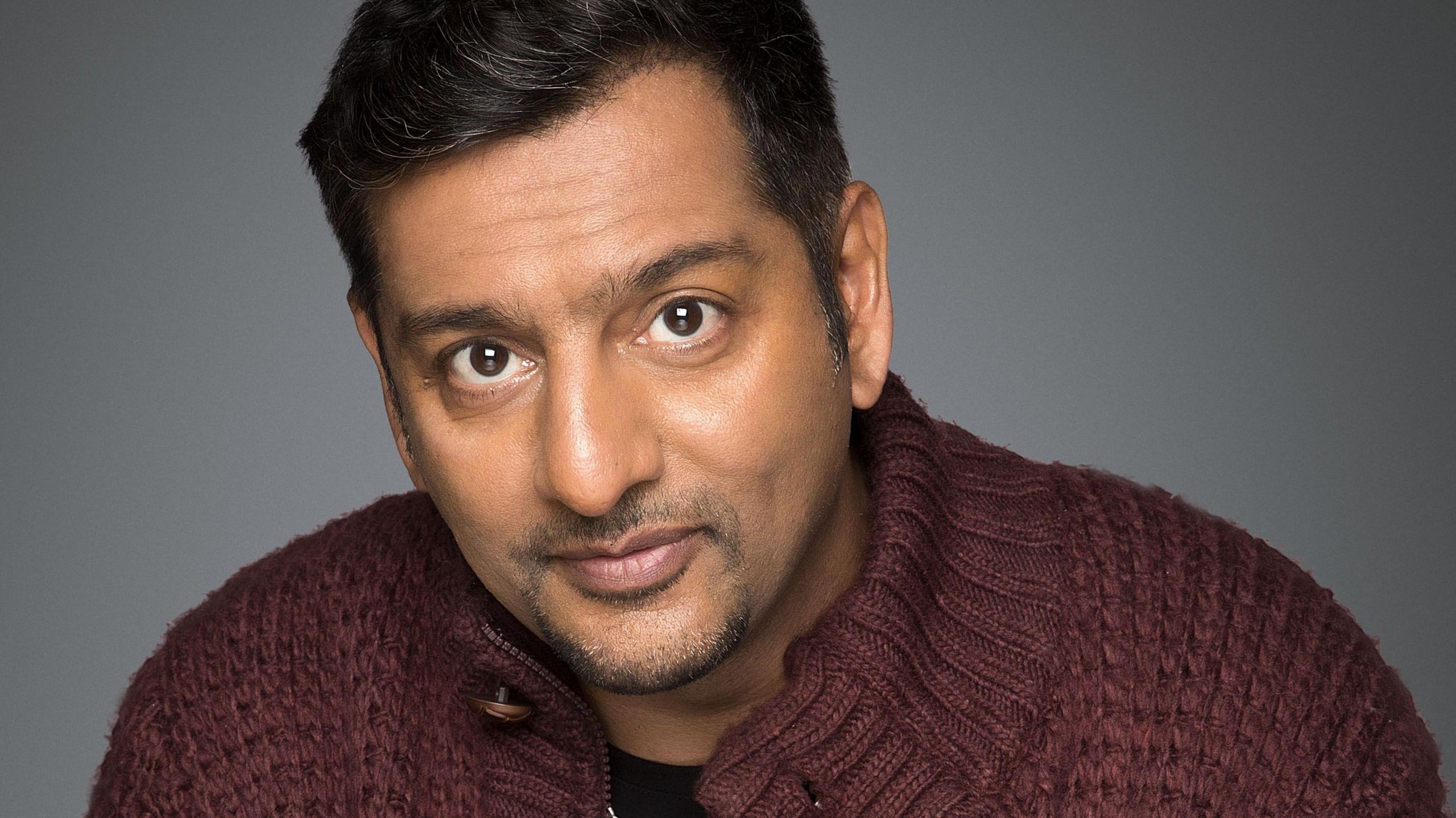 Publicity photo for Nitin Ganatra, an actor in EastEnders. He is wearing a maroon cardigan and looking at the camera with a slight smirk.