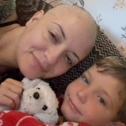 Woman sat with a boy with short brown / blonde hair. There is a toy dog and a red blanket. They are sat on a sofa with light wallpaper behind them. The woman has a shaved head.
