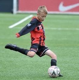 Boy with red and black football kit and short blonde hair kicking a football on a football pitch.