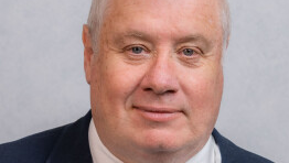 Headshot of John Wannenburgh. He wears a suit and stands against a grey background