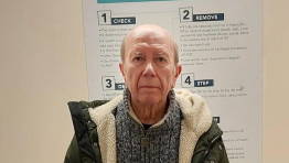 Peter Kindell at a police station: A bald man with grey hair on the side looking ahead. He is wearing a mottled grey jumper with a zip at the neck a green coat which has a white fur lining. He is standing in front of a cream coloured wall. Part of a white poster is visible on the wall. The numbers 1, 2, 3 and 4 are visible on the post plus the words "CHECK", "REMOVE", and "STEP".