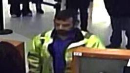 A man with short black hair and a black beard is at a cashier desk. He is wearing a hi-viz jacket and a blue shirt. 