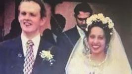 A man and a woman in wedding outfits. The man is wearing a blue suit andtie with pale flower in one lapel and the woman is wearing a headdress with flowers.