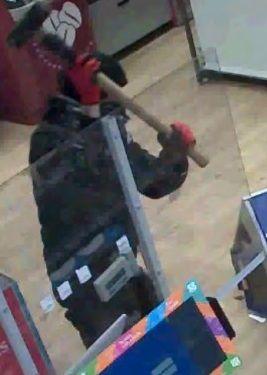 A CCTV still of a man in a black jacket, black beanie hat and red gloves wielding a sledgehammer towards a Perspex screen   