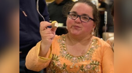 A woman with brown hair and glasses smiles. She wears a yellow flowery top and dangly earrings.