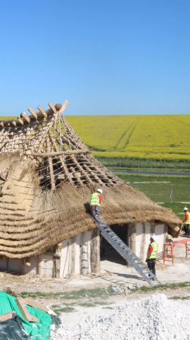 A replica neolithic hall under construction near Stonehenge with multiple volunteers in high-vis working on the structure.