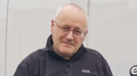 A middle aged bald man with glasses standing in front of a white van wearing a blue jumper