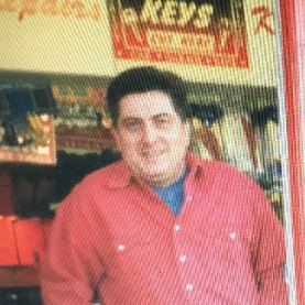 Gary Walker, pictured in an old image from 1995, outside his shop. He has dark hair and is wearing jeans and a red buttoned up shirt over a blue t-shirt, and has his hands in his pockets. He's smiling.