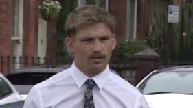 Kavan Keenan is a young man with short blonde/ brown hair and a moustache. He is walking towards the gates into the court as the picture is taken. He is wearing a white long sleeve shirt and a black patterned tie. Behind him is a silver car and two white cars parked along the street in front of red brick terrace houses.